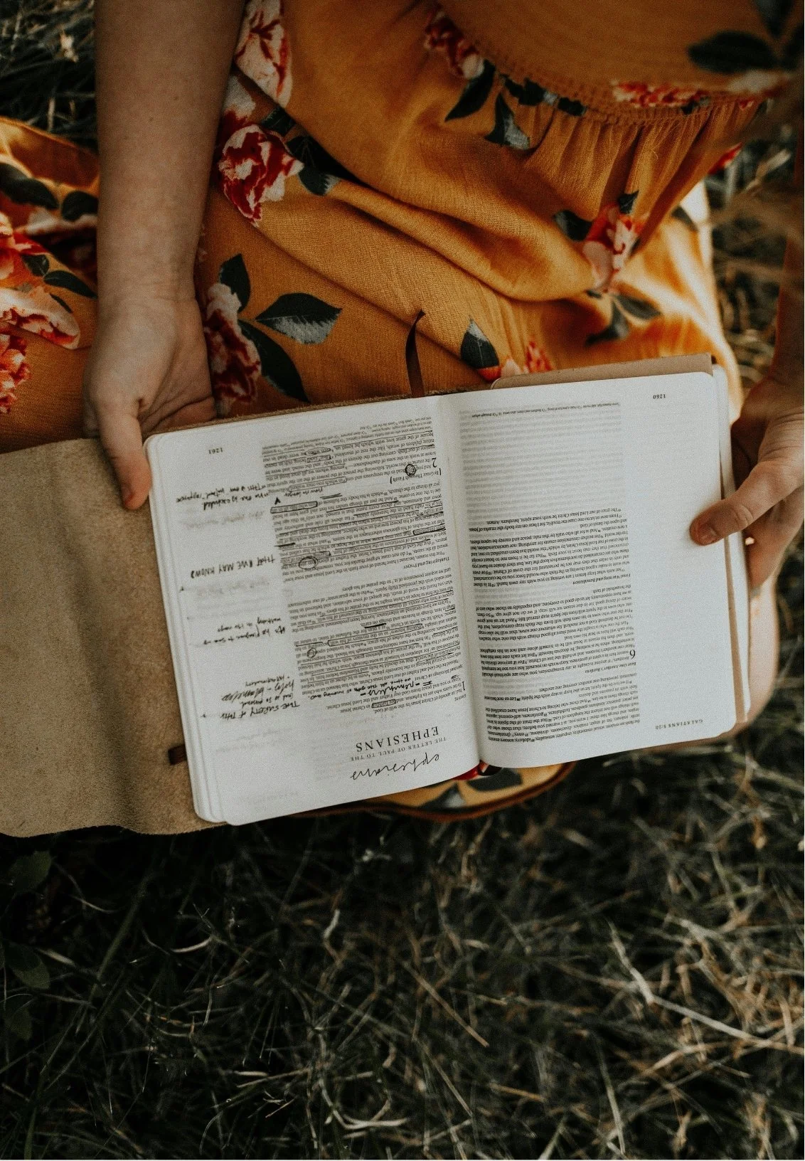 Person sitting outdoors, holding an open Bible with handwritten notes, wearing a yellow floral dress.