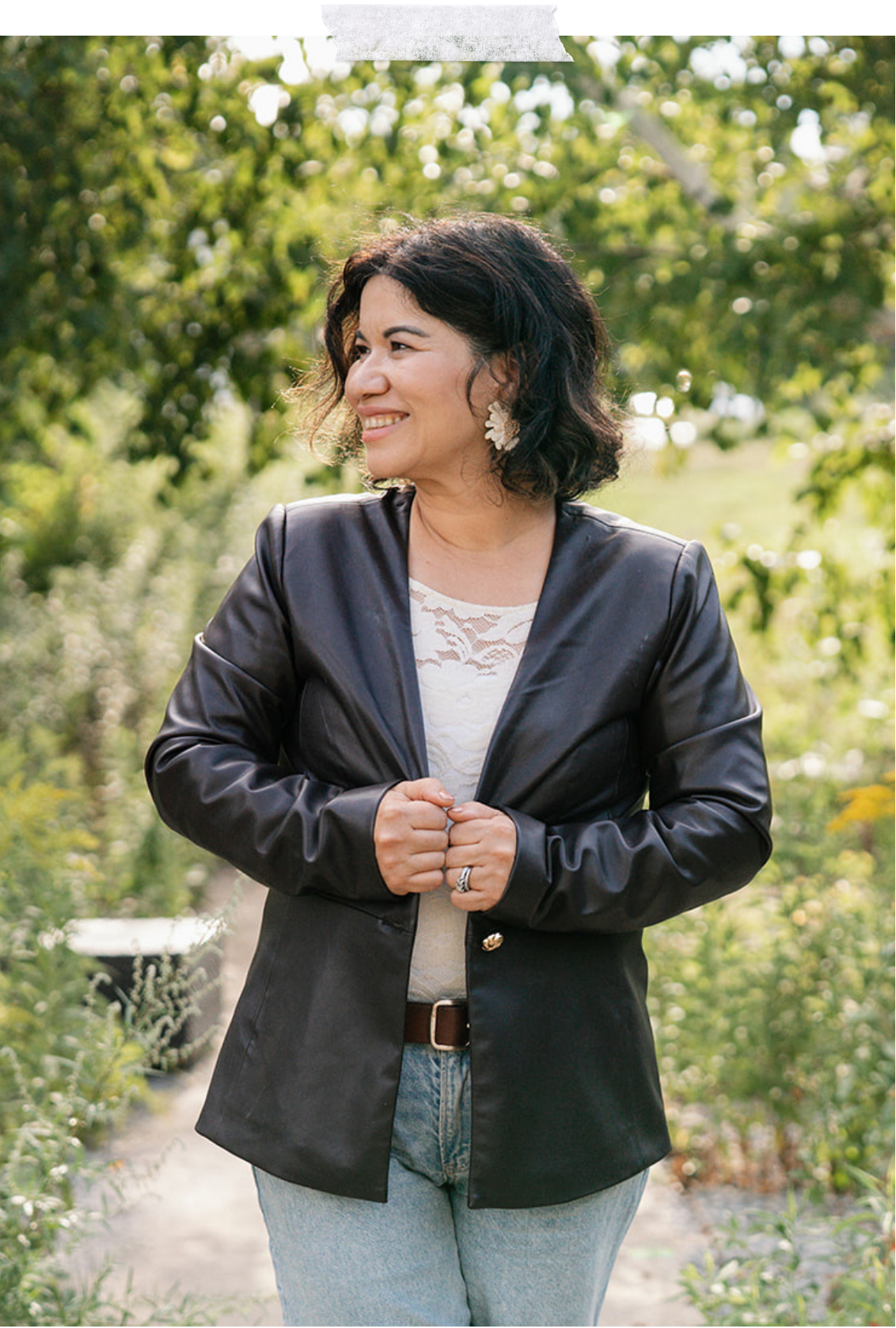 Boston-based therapist Adaris Pickett wears a black leather jacket, white lace top, and light blue jeans, standing outdoors among green trees and plants.