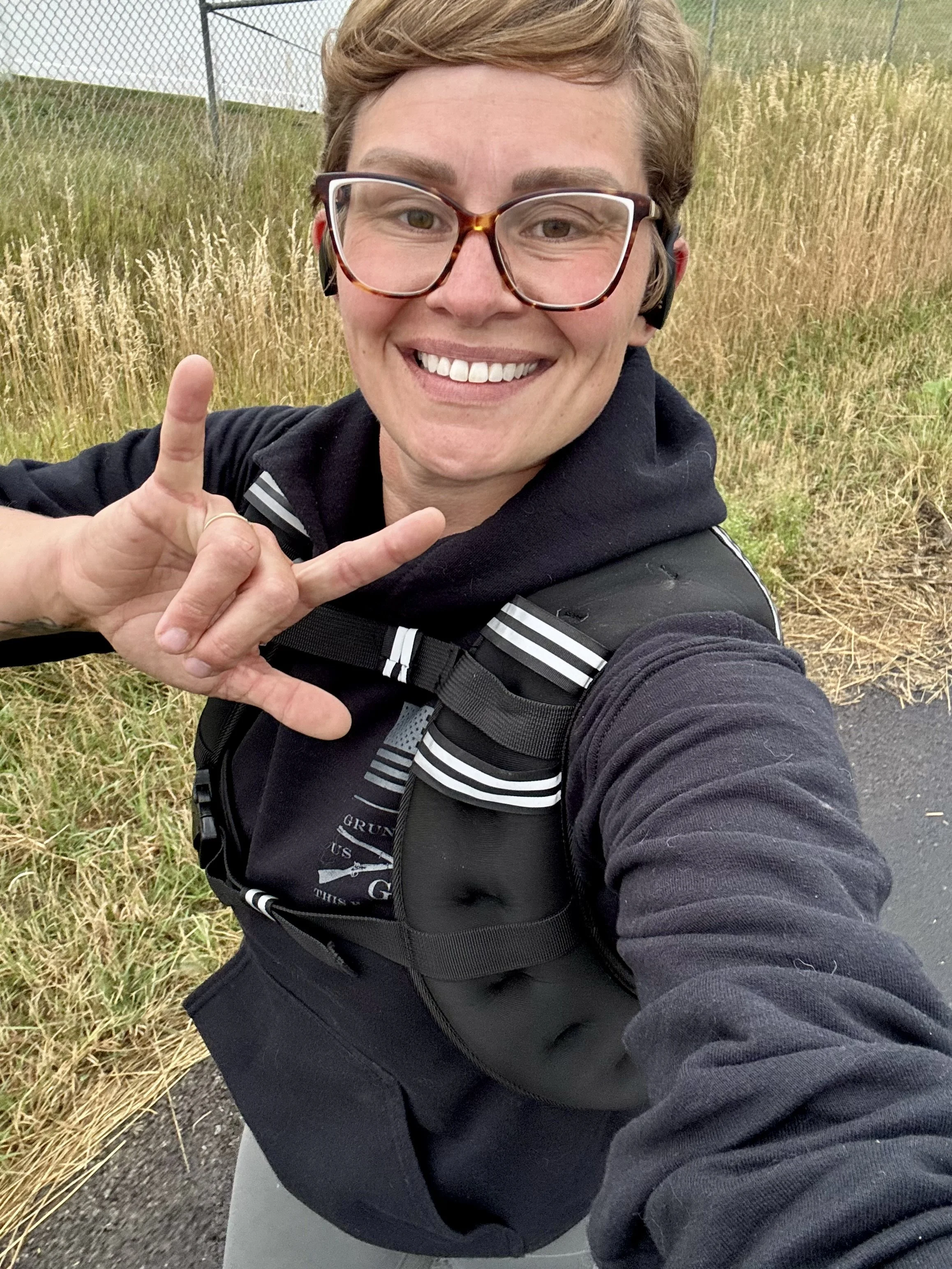A smiling person taking a selfie outdoors near a grassy field, wearing glasses, a black hoodie, and a backpack.