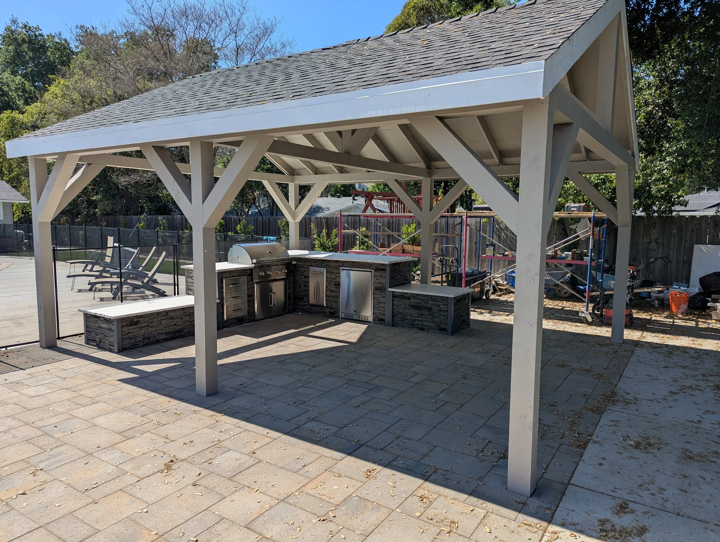 An outdoor cooking area with a wooden pavilion, stone countertops, a grill, and outdoor seating, surrounded by a fenced backyard with trees and construction equipment in the background.