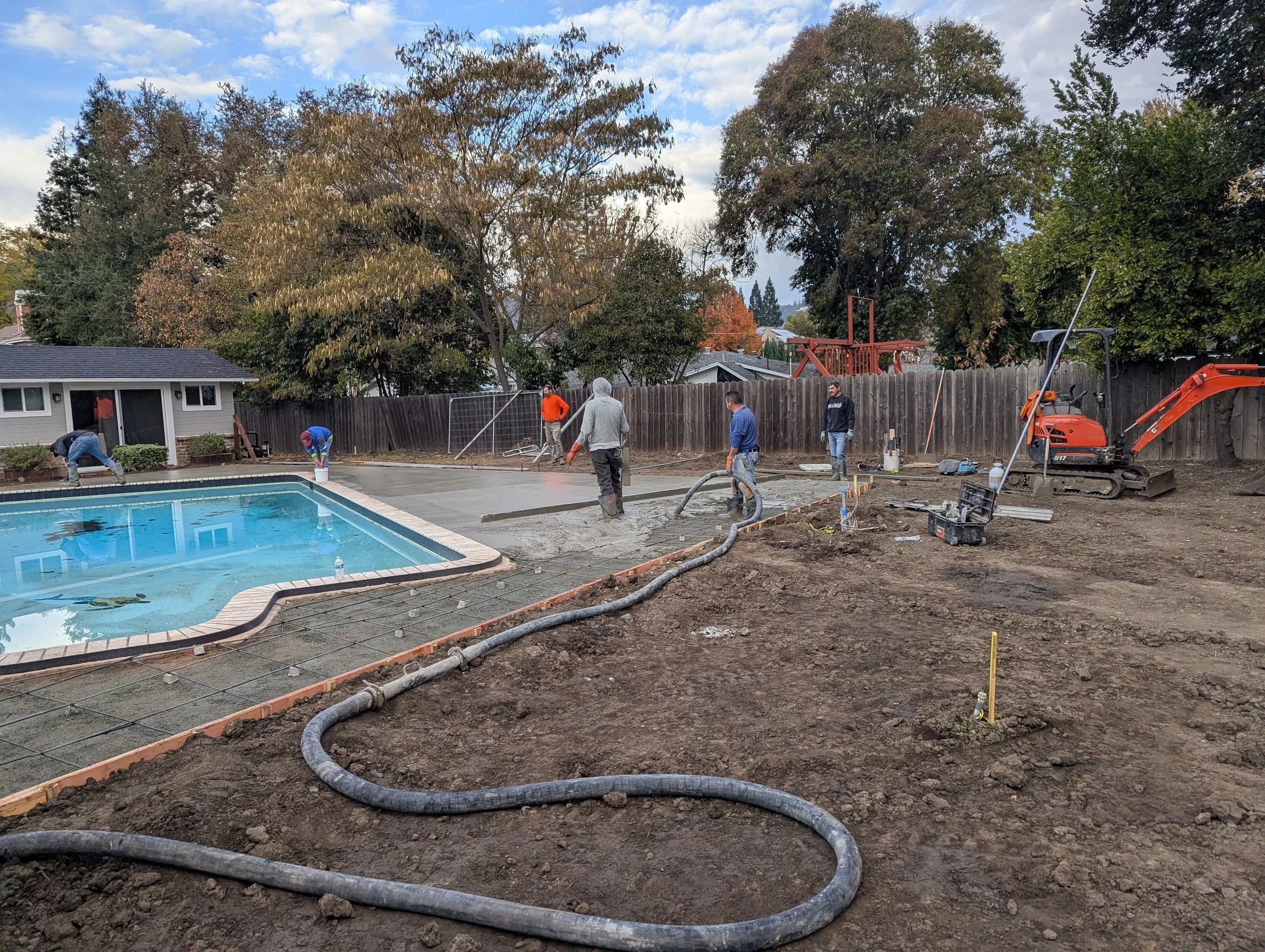 Workers pouring concrete for a backyard pool renovation, with construction equipment and tools around, and a fenced yard with trees in the background.