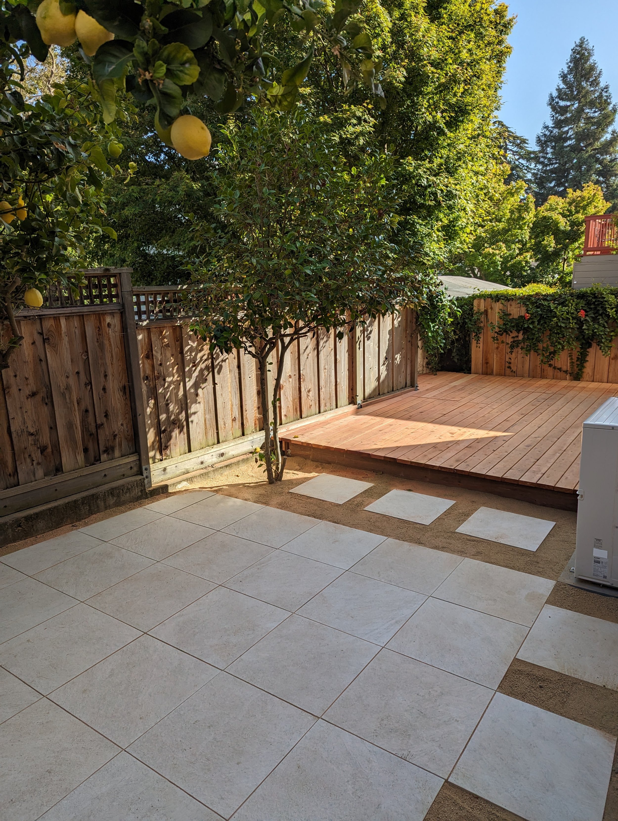 Backyard patio with tiled area, wooden deck, tree, and fence, with trees and sky in the background.