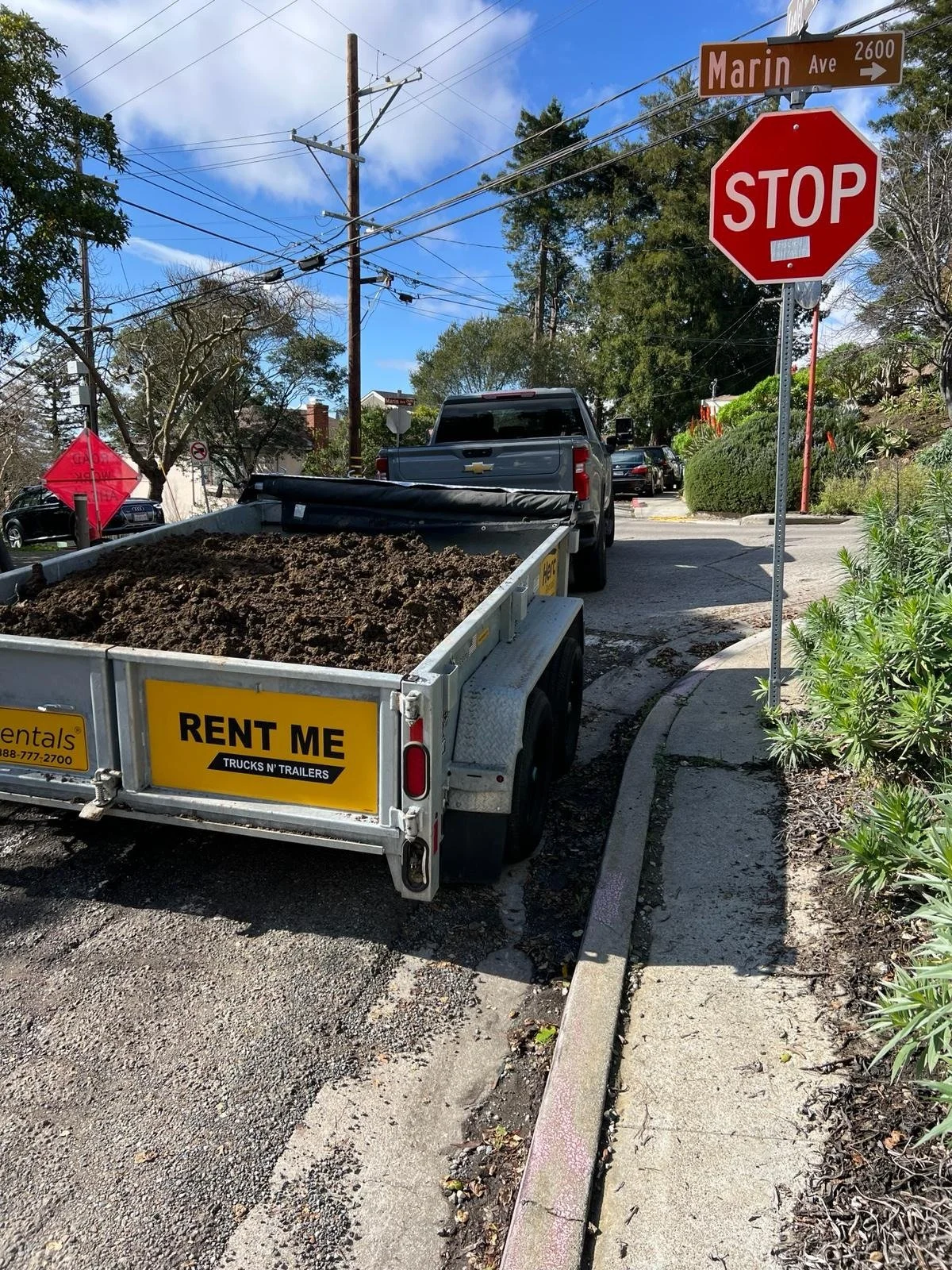 A street corner with a stop sign, a street sign reading Marin Ave, and a trailer filled with dirt parked on the side of the road. There are trees and utility poles with power lines in the background.