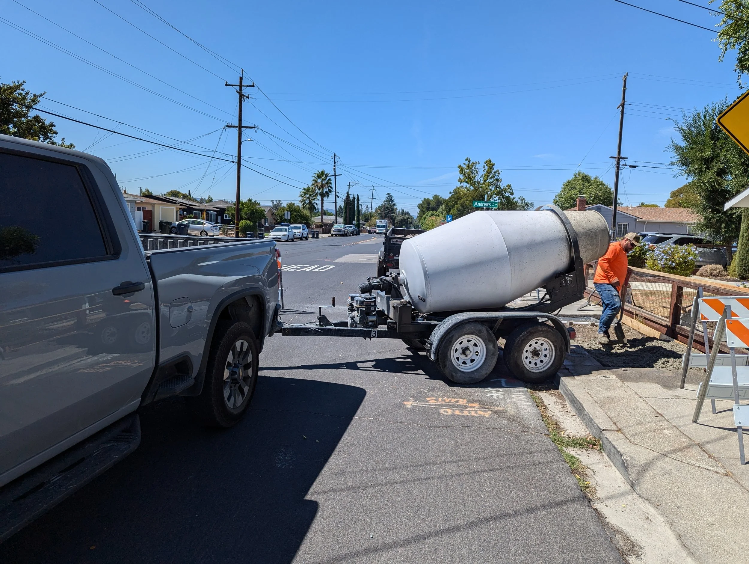 A construction site on a city street showing a large concrete mixer truck on a trailer, a worker in an orange shirt and jeans working with shovels, and parked cars along the street.