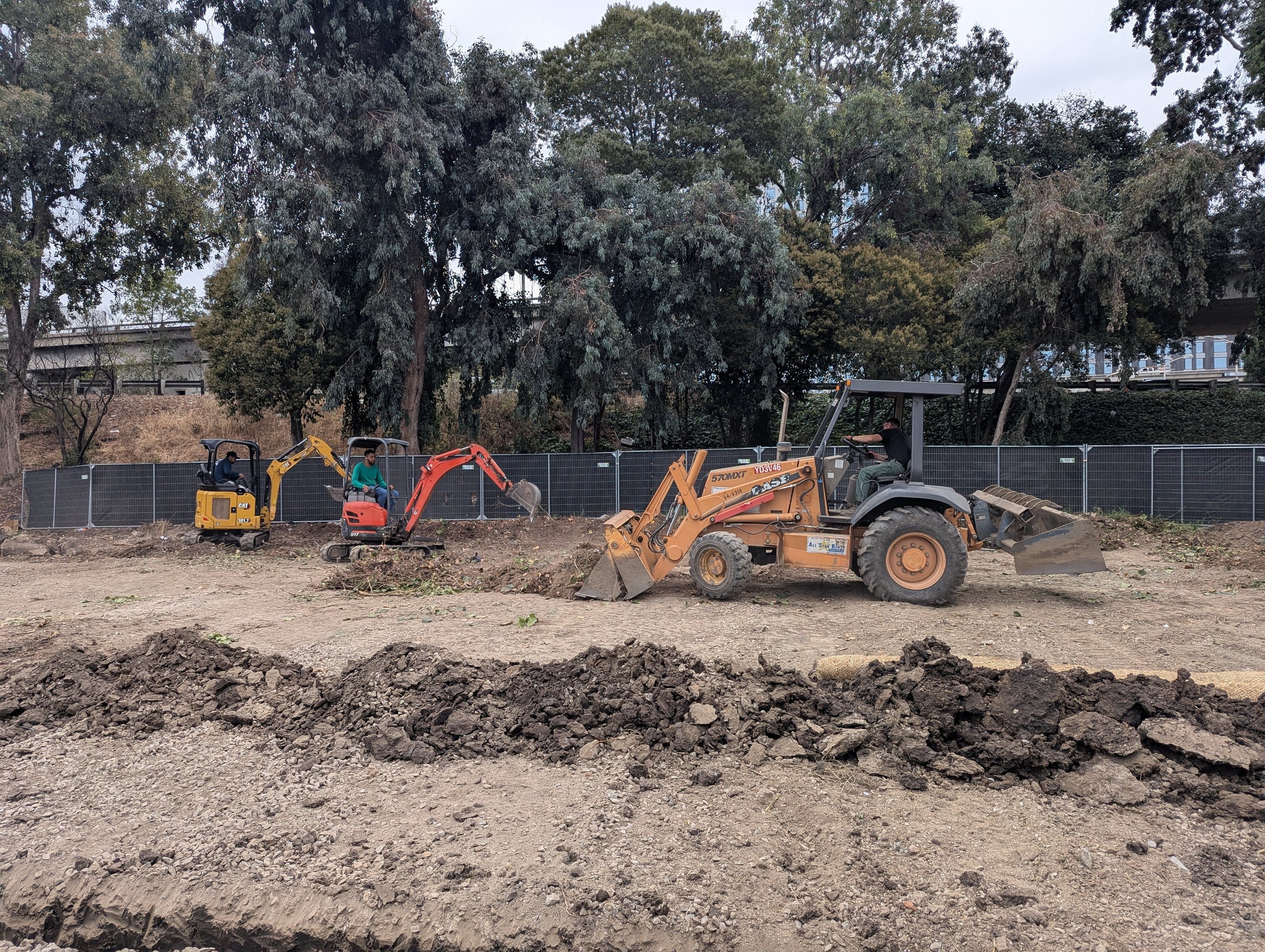 Construction site with three workers operating small excavators and a front loader, digging and moving soil, with trees and a fence in the background.