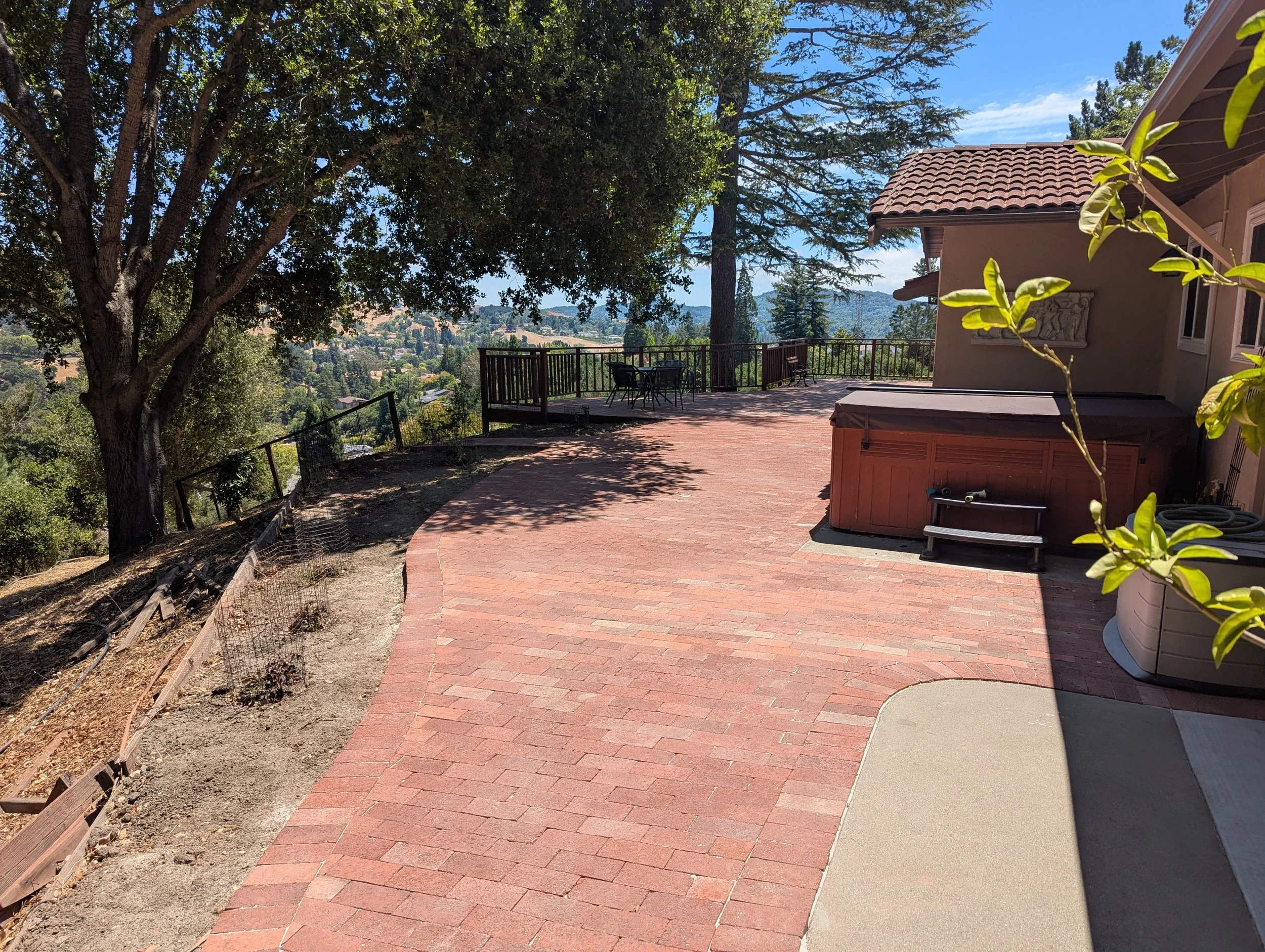 A spacious outdoor patio with brick flooring and a hot tub, overlooking a scenic view of trees and distant hills under a bright blue sky, partially shaded by a large tree.