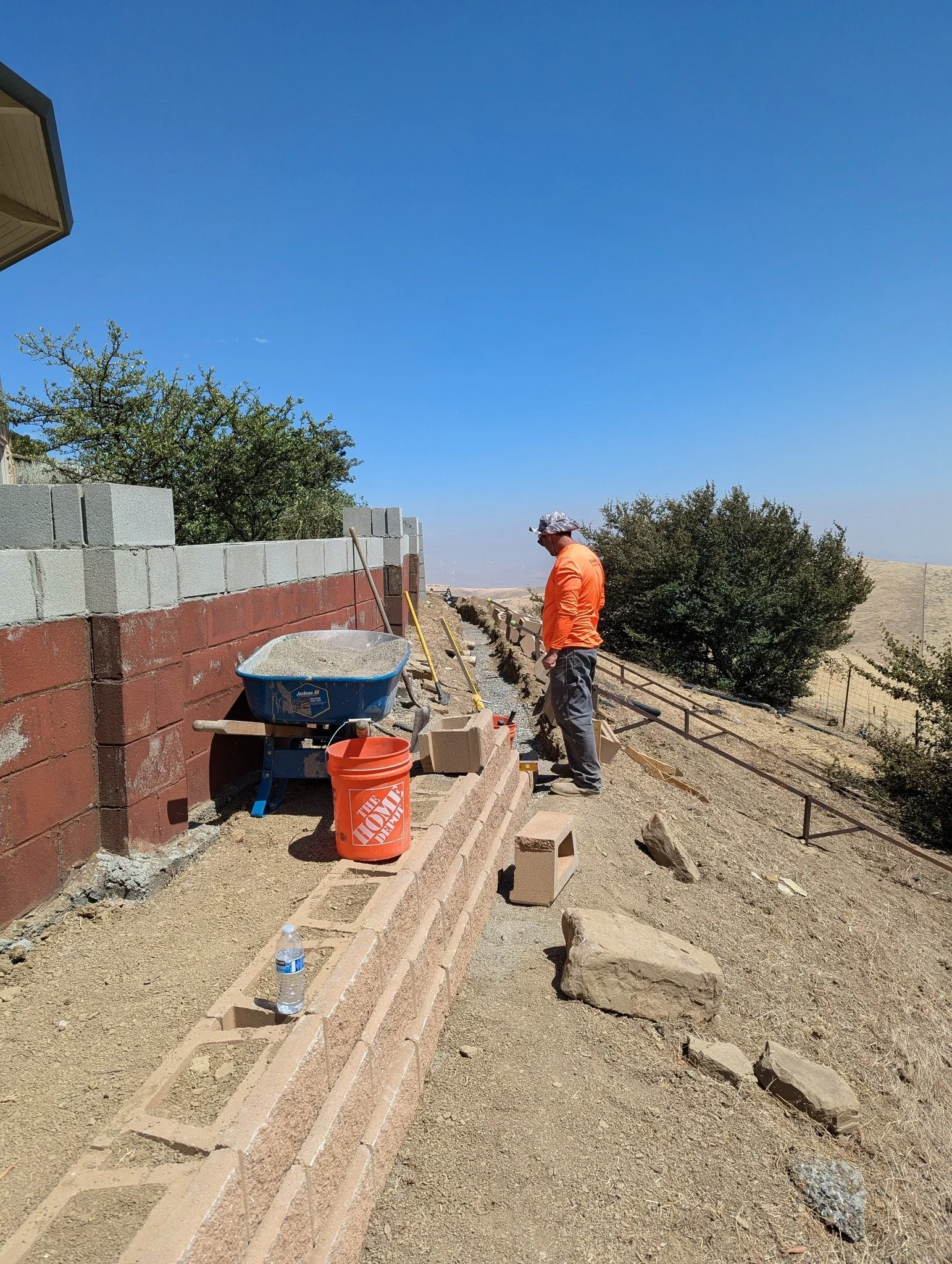A construction worker in an orange shirt and gray pants working on building a brick wall on a barren hillside. Construction tools and materials, including a wheelbarrow, an orange bucket, and bricks, are nearby. The background features a clear blue s