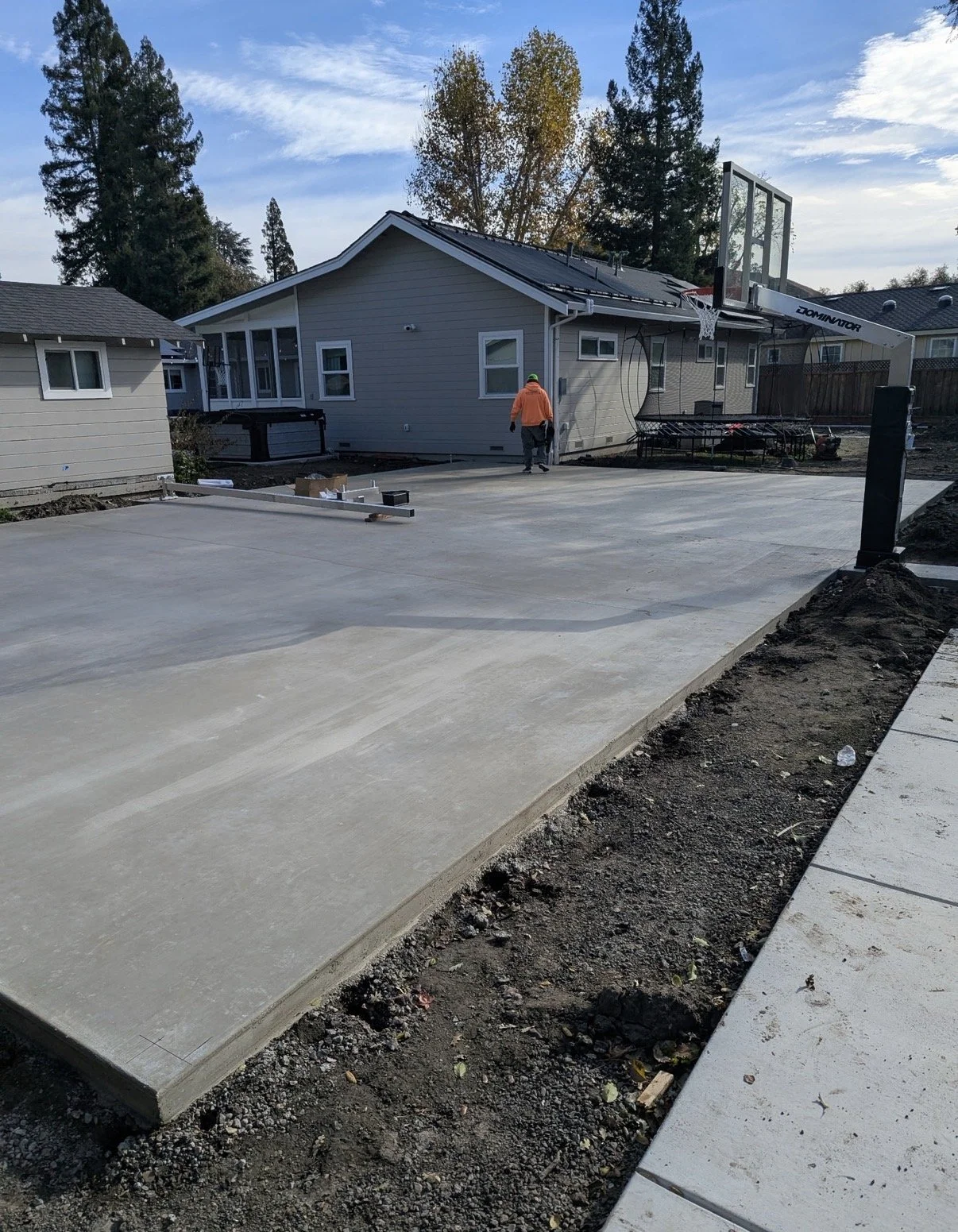 A newly poured concrete driveway in a residential backyard with a basketball hoop, a man walking, and houses in the background under a partly cloudy sky.