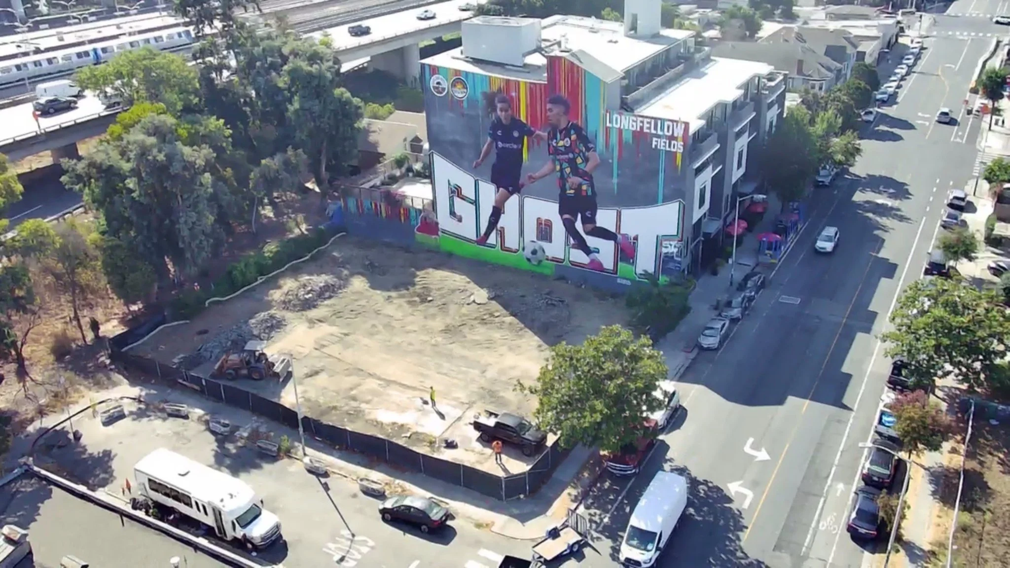 A large mural on the side of a building featuring two soccer players, with a colorful background and the words "Longfellow Fields." The mural is in an urban area with nearby streets, parked cars, and trees.