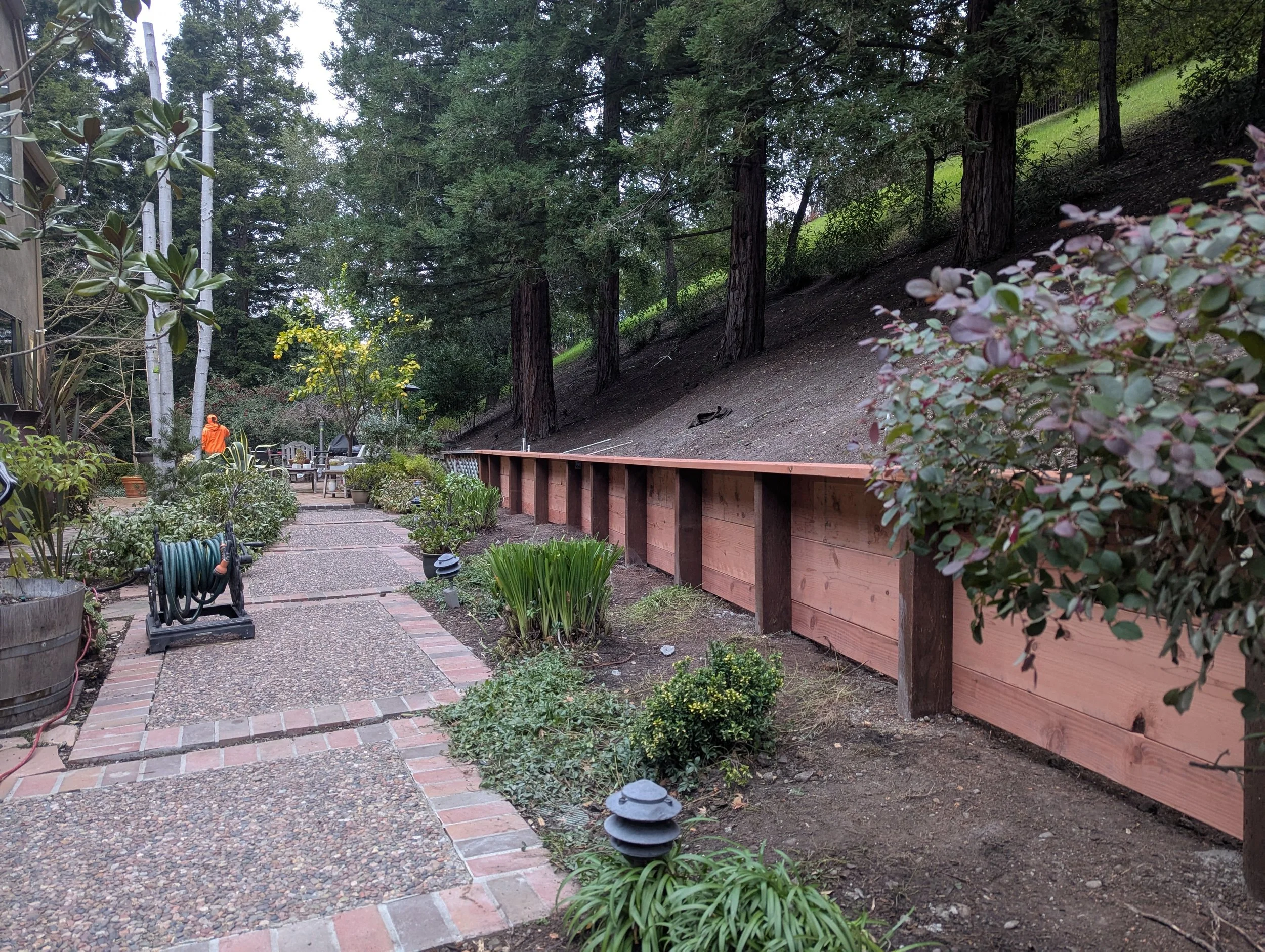 A landscaped backyard garden with a paved walkway, potted plants, garden hose, and outdoor seating. A wooden retaining wall supports a hillside with trees. A worker in orange overalls is seen in the distance.