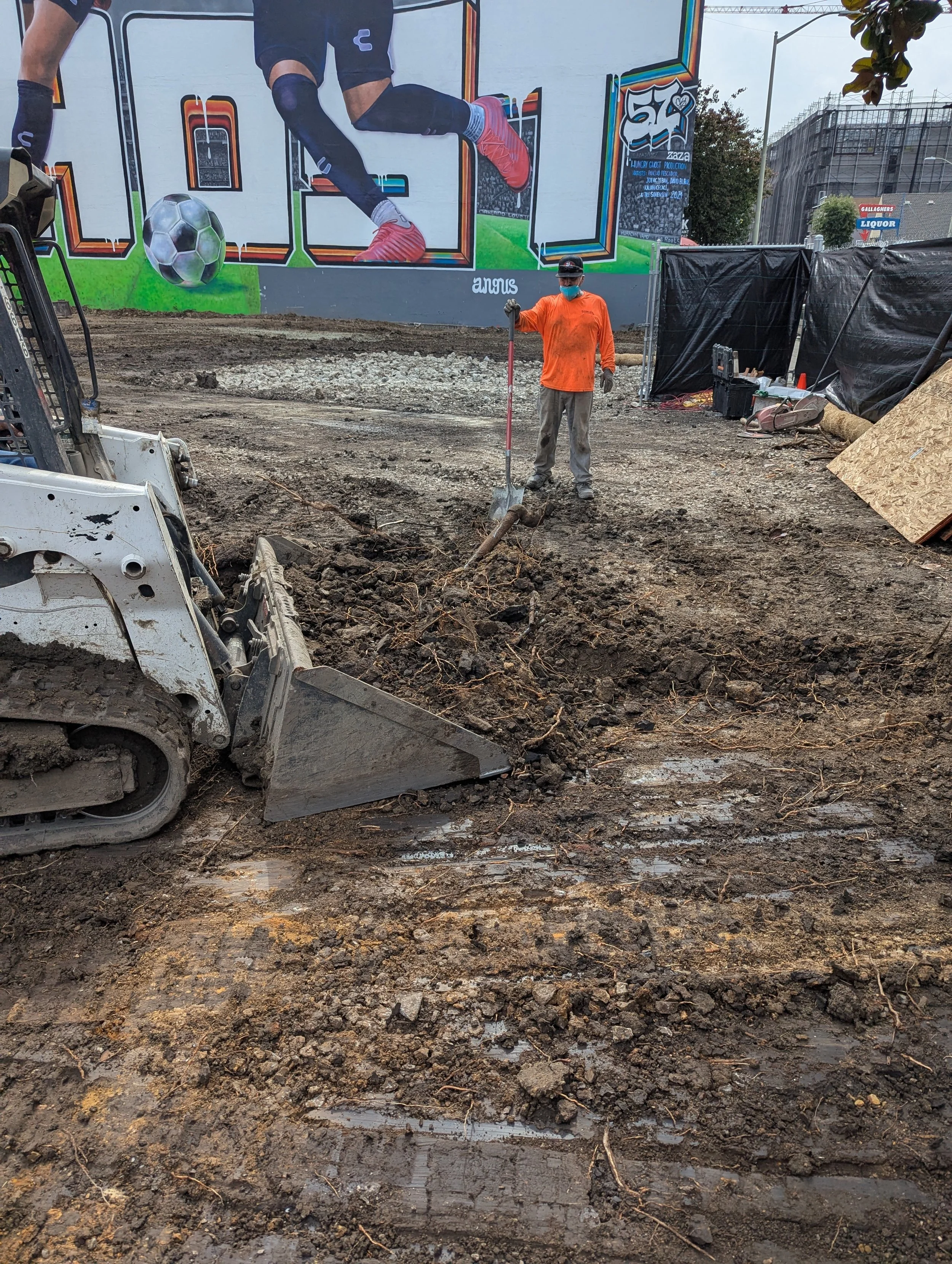 A construction worker in an orange shirt, gray pants, and a face mask stands with a shovel on a dirt site, with a small excavator in the foreground. There is a large colorful mural on the back wall featuring a soccer player and a soccer ball.