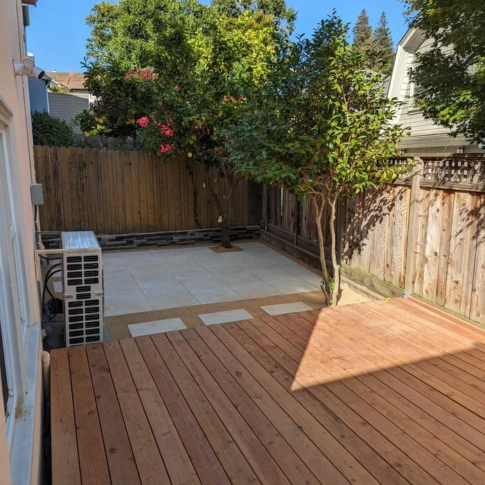 A backyard with a wooden deck, a small tiled patio area, and two trees along a wooden fence. An outdoor air conditioning unit is on the left side.