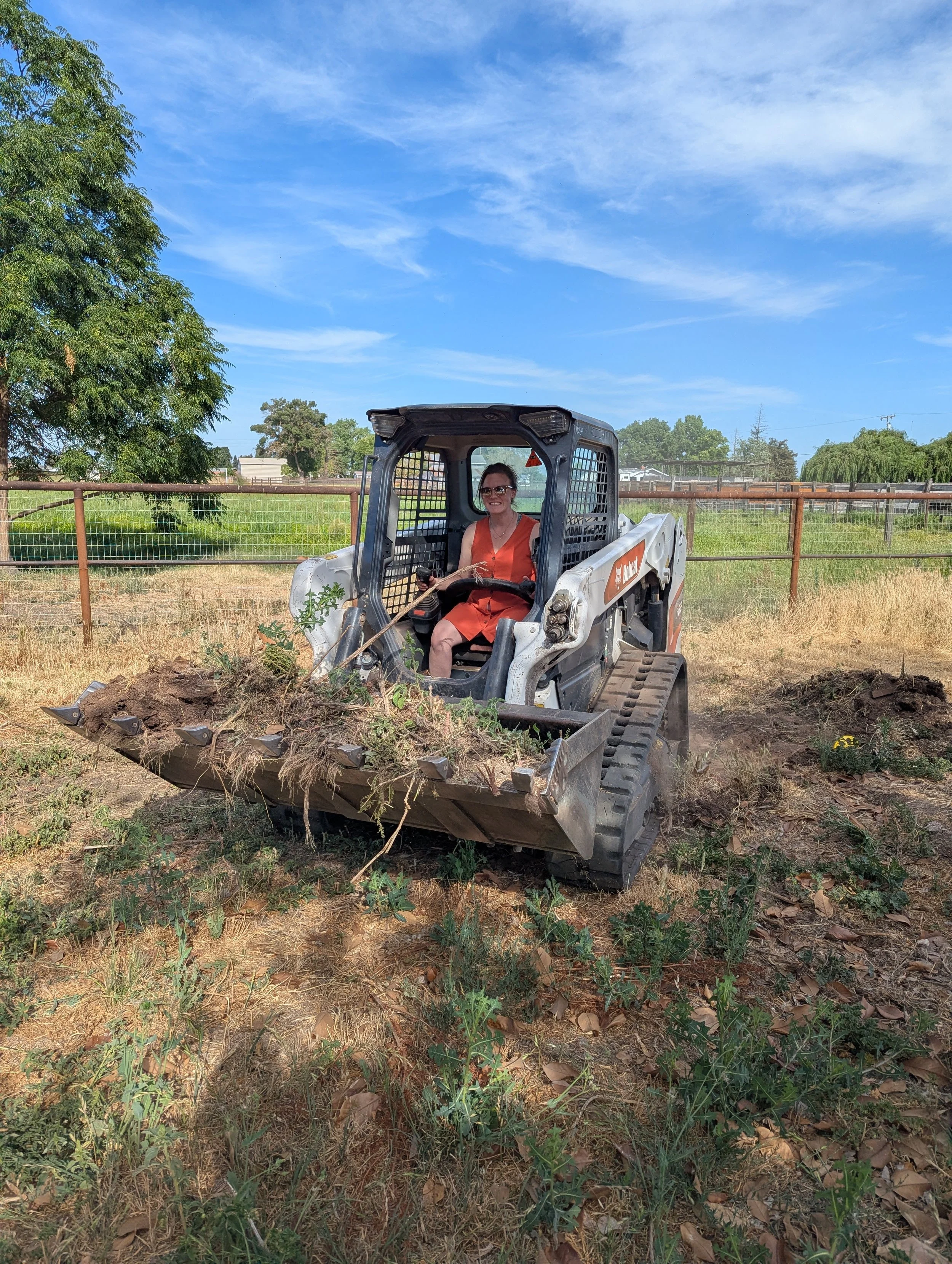Woman in orange dress sitting in a skid steer loader with dirt and plants in its bucket, on a dry, grassy field with a fence and trees in the background under a blue sky.