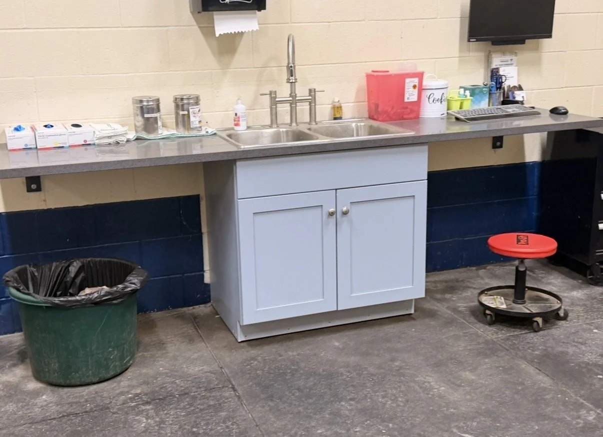 A kitchen or break room area with a gray countertop, sink, and white cabinet. Items on the counter include paper towels, cleaning supplies, a pink bin, a coffee canister, and a computer keyboard and mouse. There is a green trash bin with a black plas