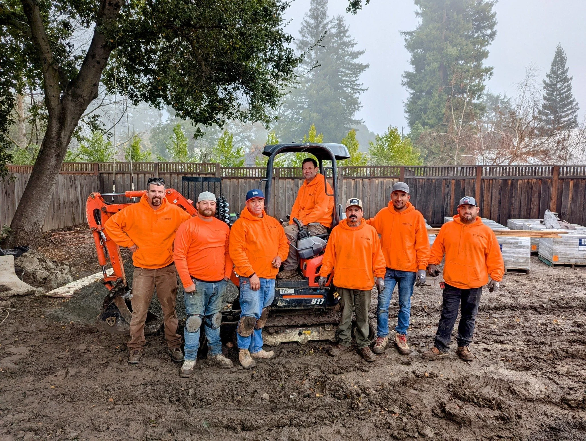 Group of seven construction workers in orange jackets standing outdoors on muddy ground, with a small excavator, trees, and a wooden fence in the background.