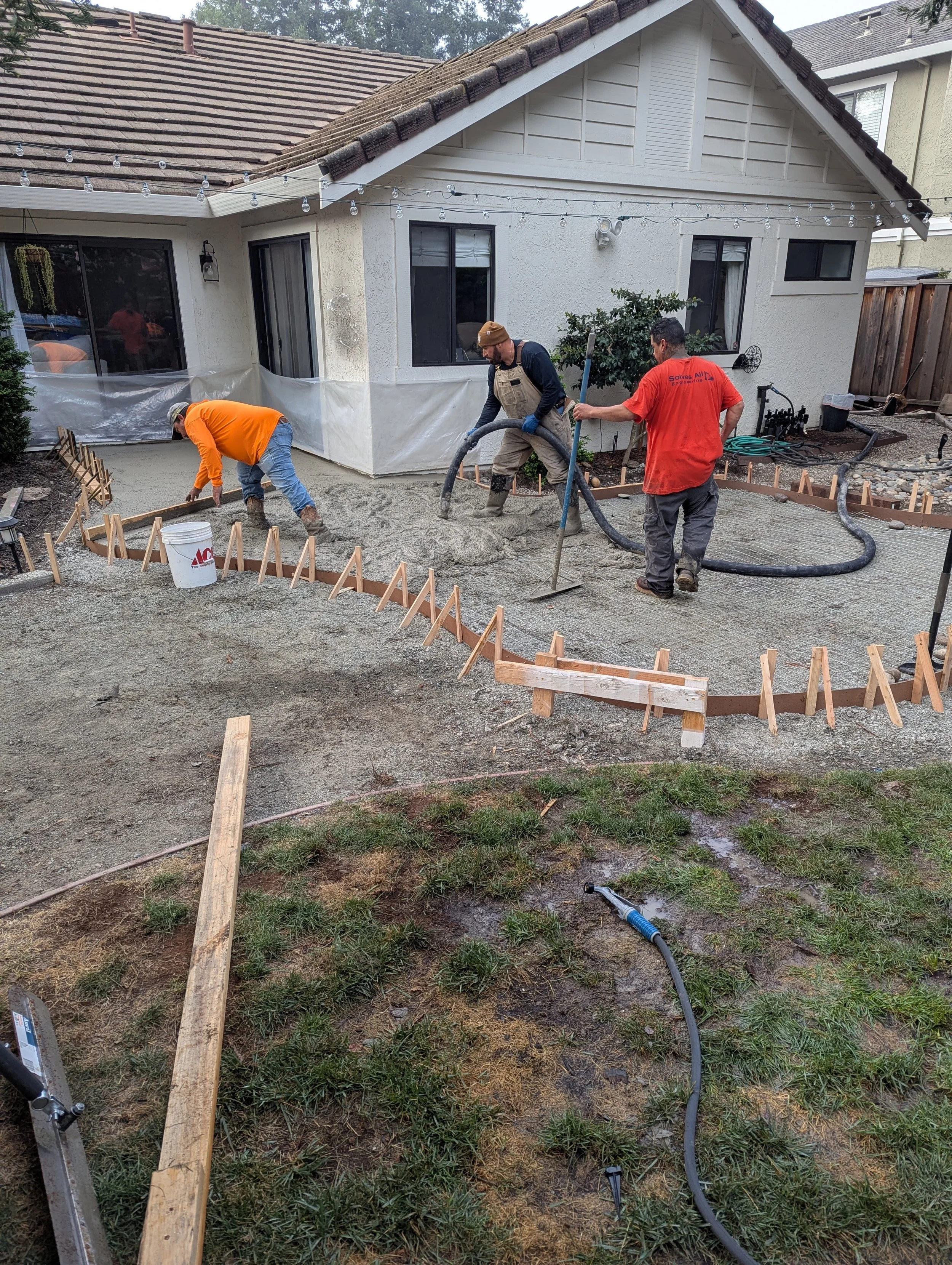 Three workers are pouring and leveling concrete in a backyard during a construction project. They are surrounded by wooden forms, with a house and string lights in the background.