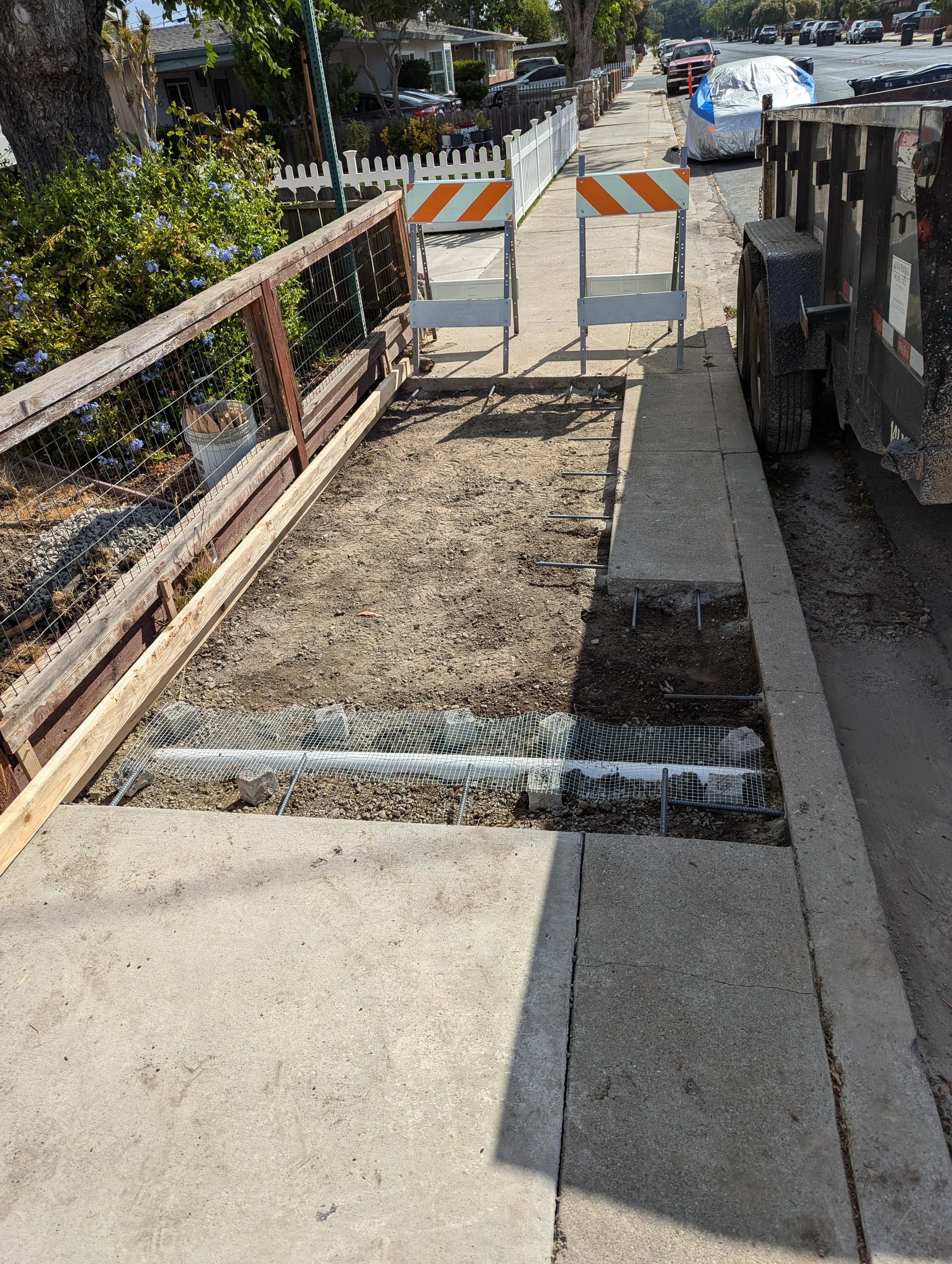 Sidewalk with construction work in progress, fenced-off area with dirt, and barriers blocking part of the sidewalk in a residential neighborhood.