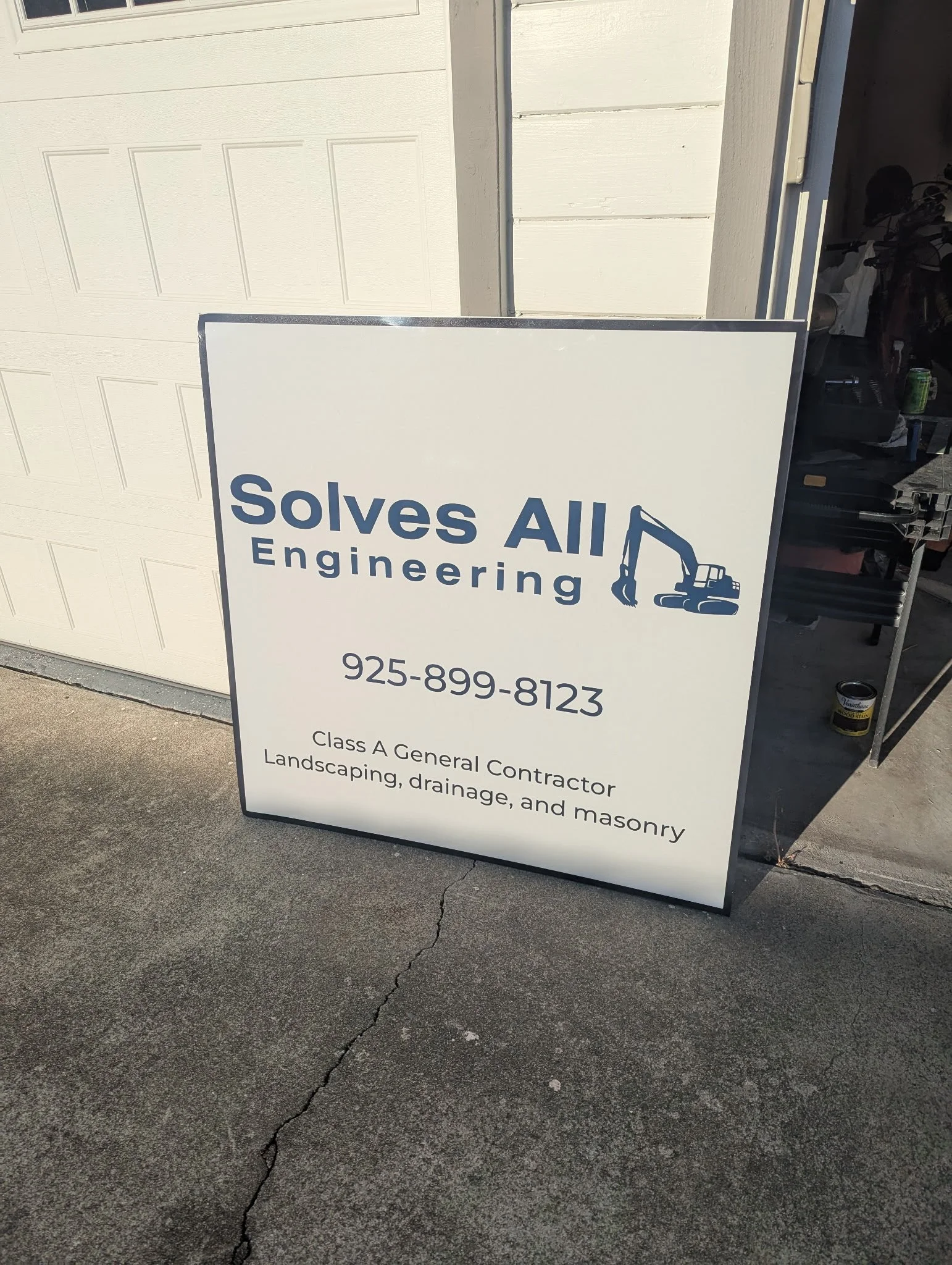 A white signboard for Solves All Engineering placed on a concrete driveway outside a garage. The sign features a blue logo with an excavator illustration, the company's phone number, and services including general contracting, landscaping, drainage, 