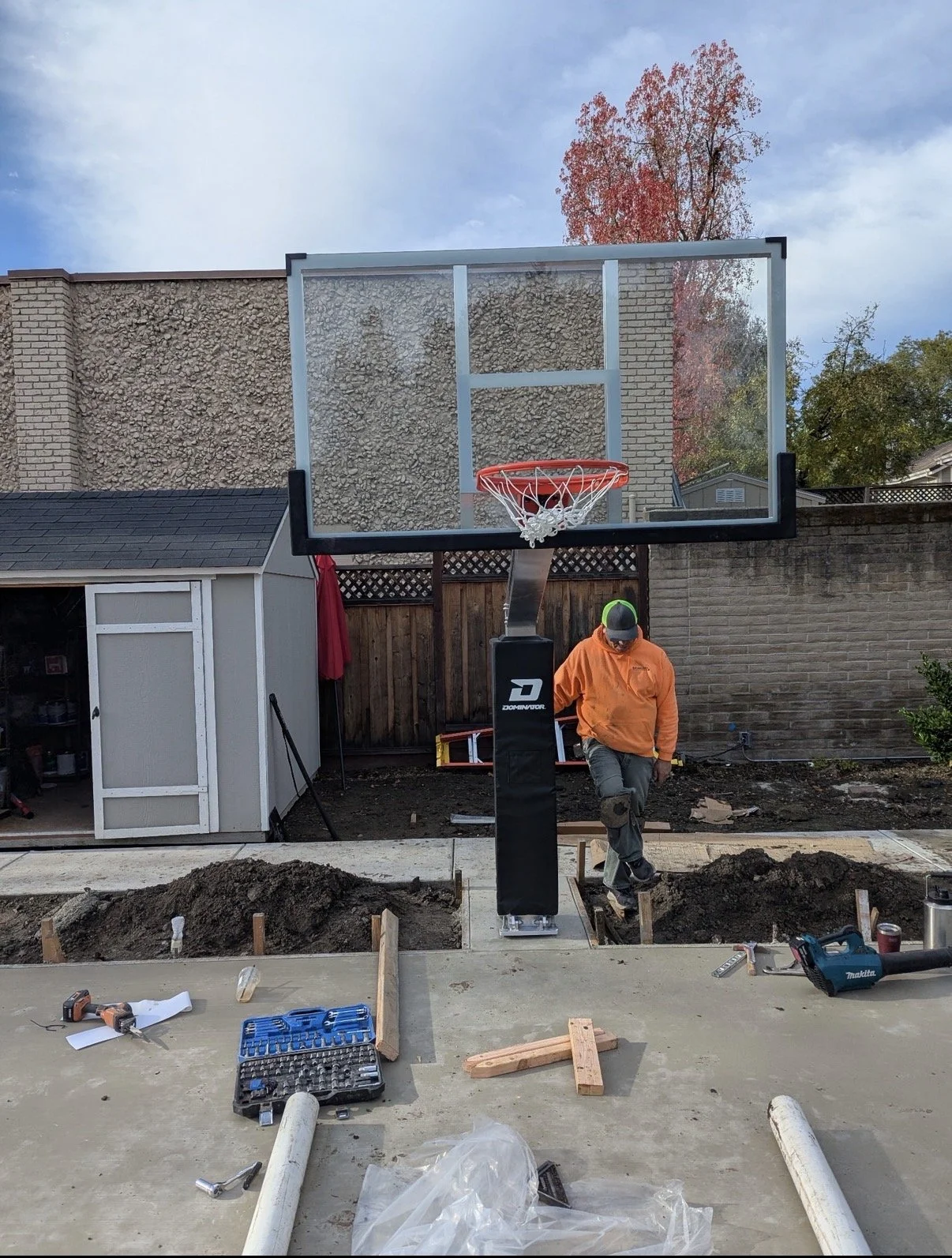 Person installing a basketball hoop in a backyard with a concrete surface, various tools and construction materials scattered around, and a shed in the background.