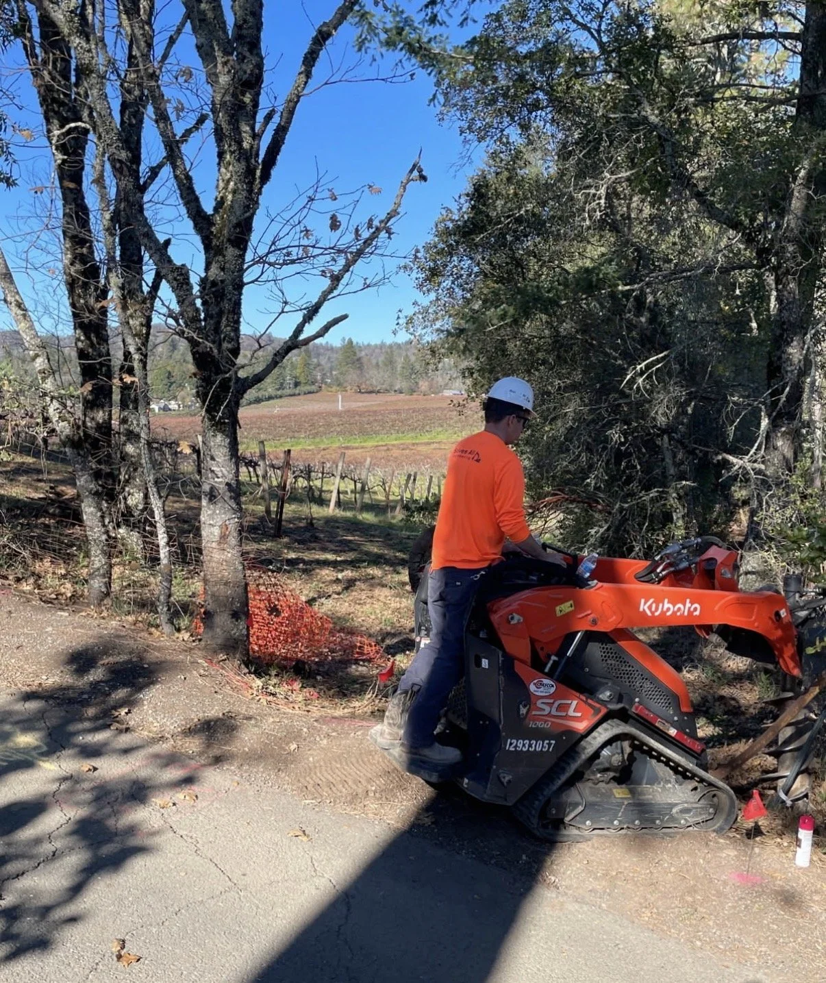 A person wearing an orange long sleeve shirt, jeans, and a white helmet operating a small Kubota excavator on the side of a rural road, with trees and farmland in the background.