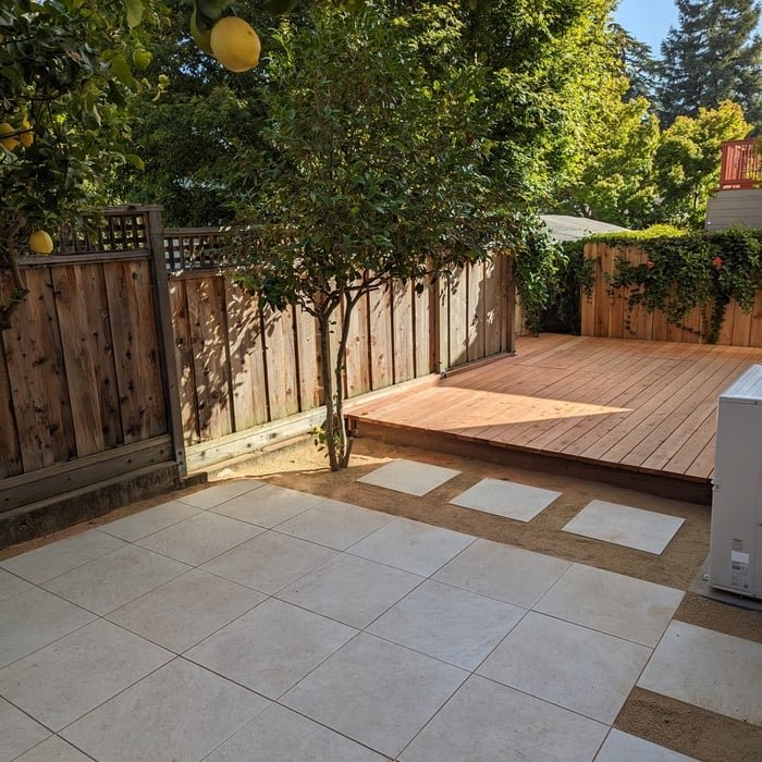 A backyard with a tiled patio, a small wooden deck, a tree, and a fenced yard with green foliage.