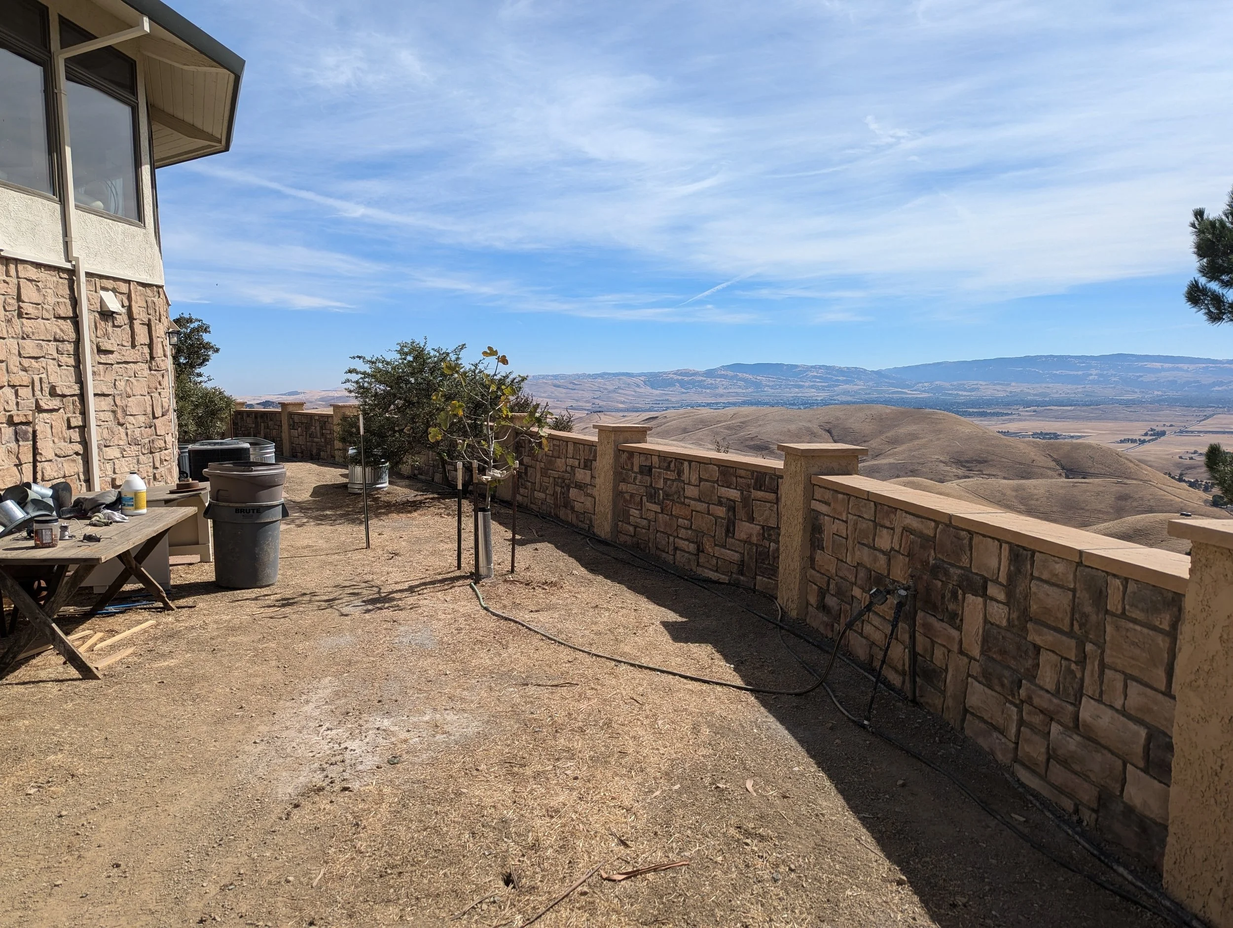 An outdoor patio area with a stone wall railing overlooking a hilly landscape. There's a wooden table, trash can, and small trees planted along the edge, against a house with beige and white exterior walls. The sky is partly cloudy.
