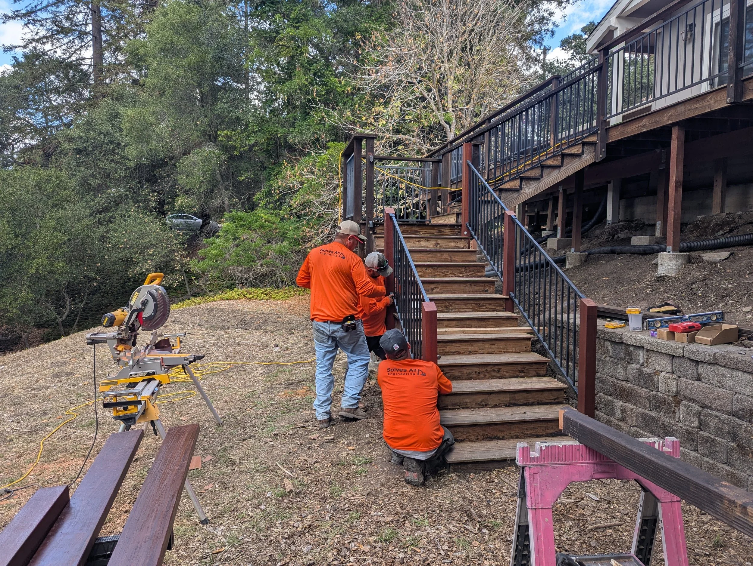 Four workers in orange shirts are installing a staircase with wooden steps and black metal railings outside a house, working on assembly and measurements.