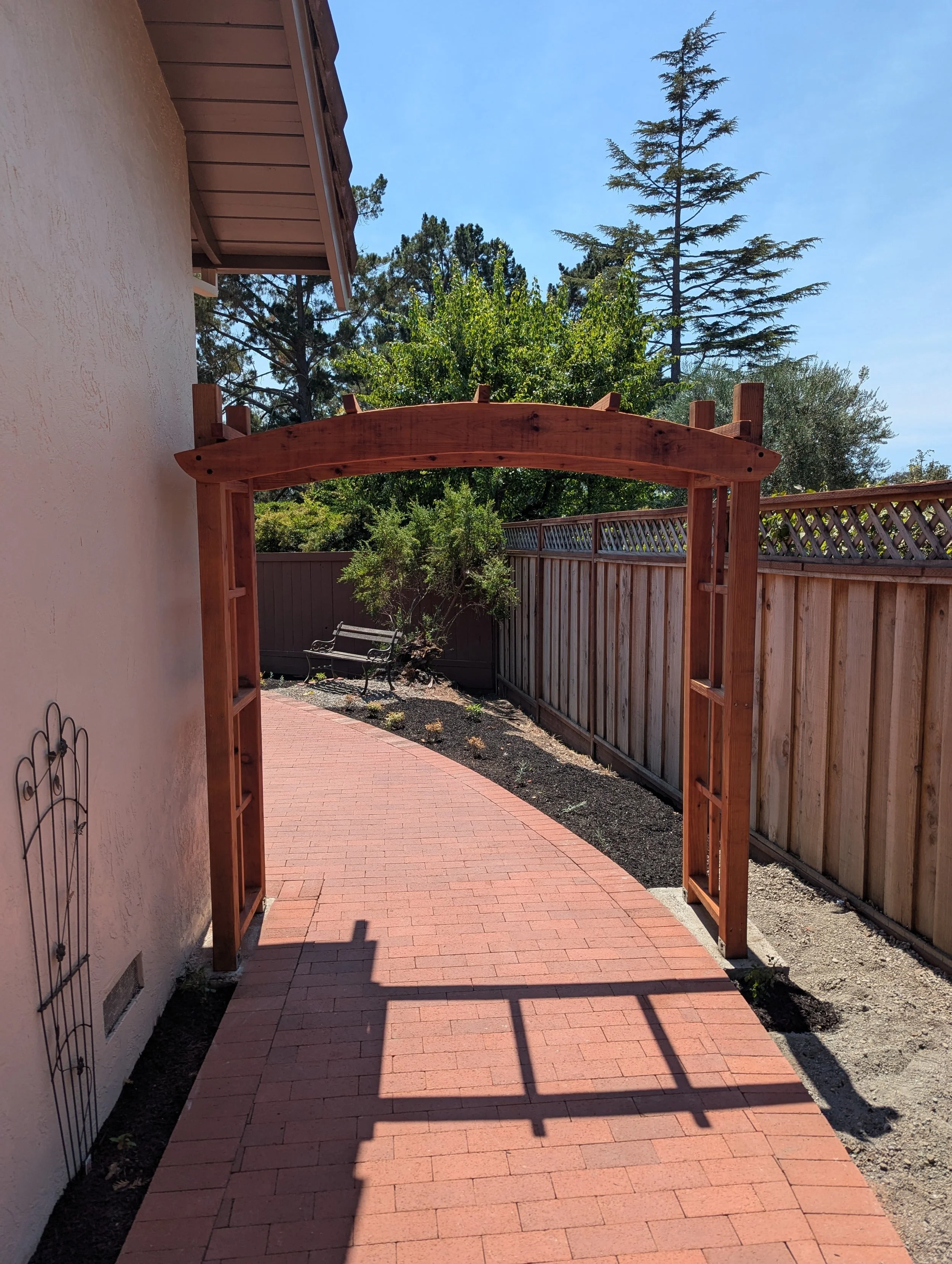 Wooden garden arch over a brick pathway, with trees and a bench in the background, and a wooden fence on the right.