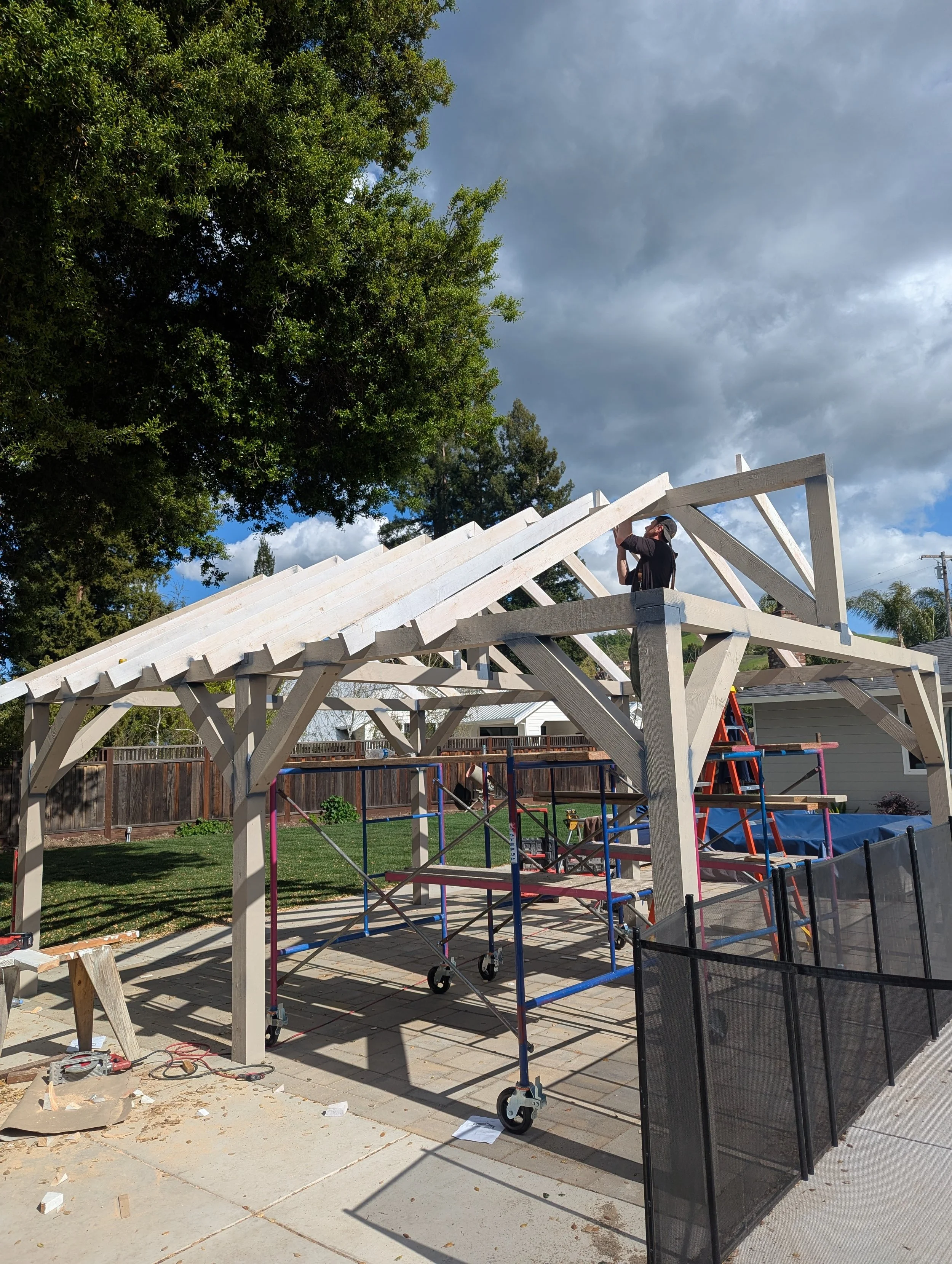 A person working on a wooden pergola frame in a backyard, with scaffolding and a ladder, clouds in the sky, trees, and a house in the background.