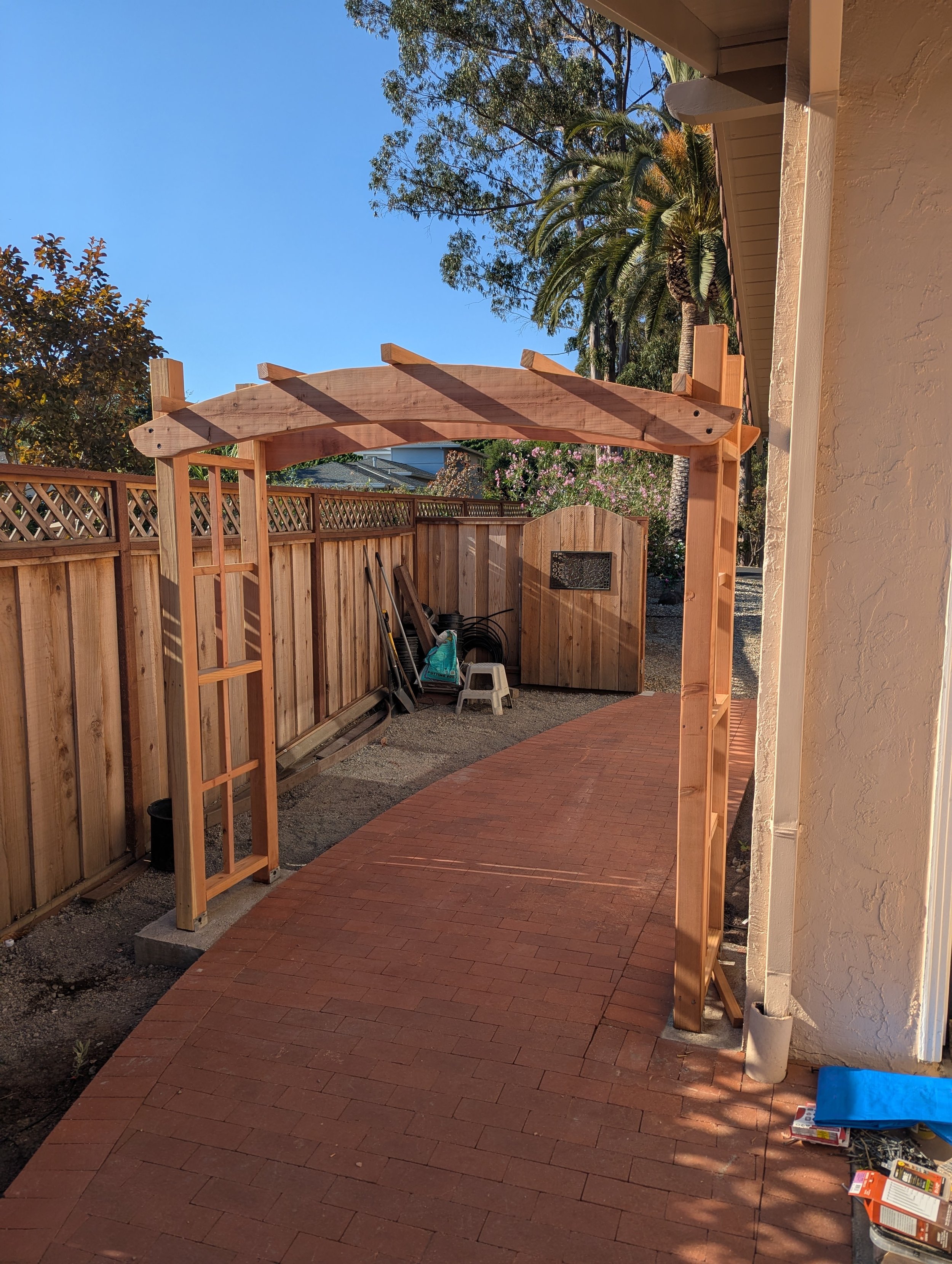 Wooden arch framing a garden pathway with a private backyard fence and trees in the background.