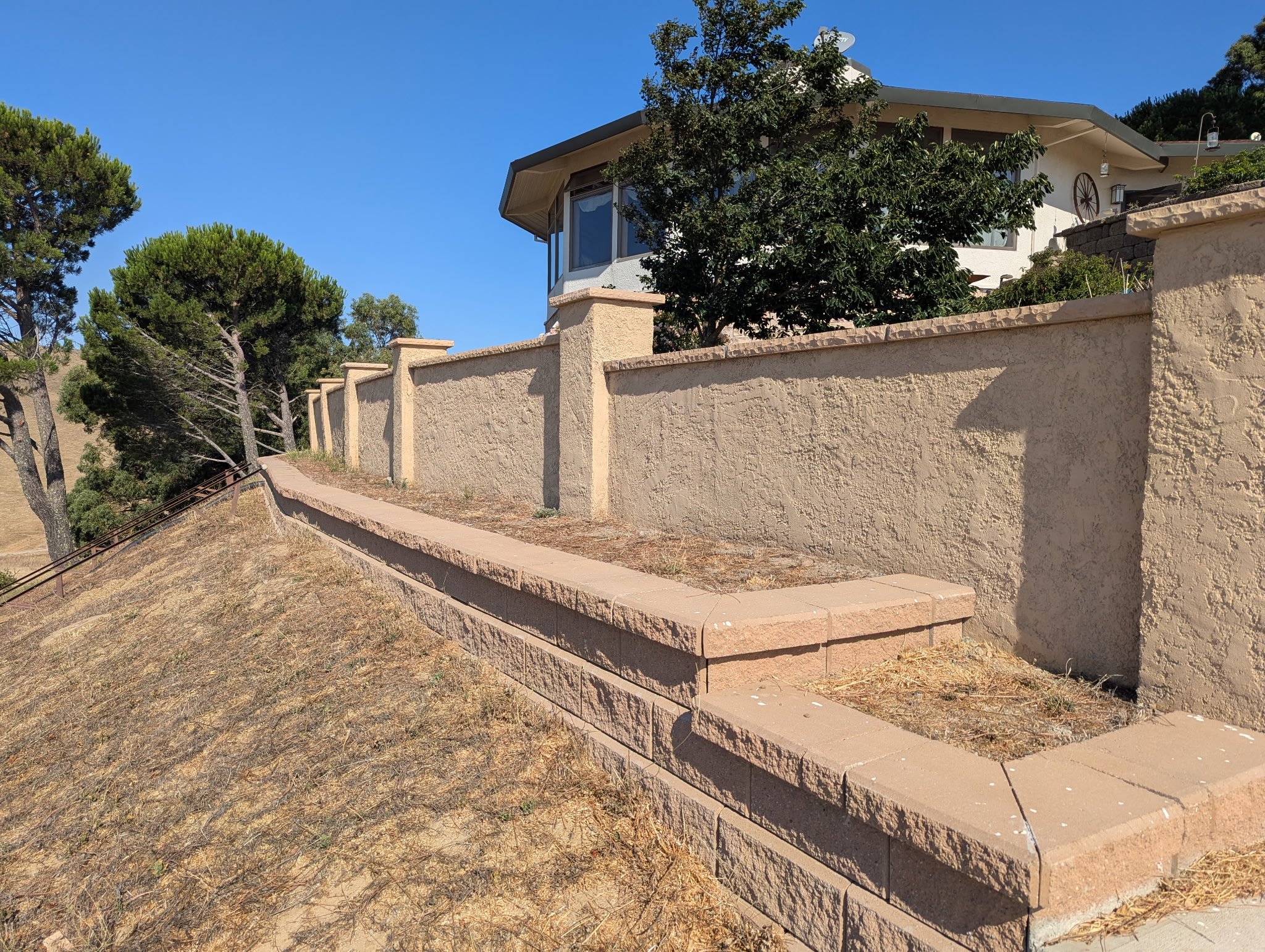 A beige stucco wall with a curved top and a brick retaining wall beneath it, situated on a sloped dirt ground with some dry grass, and a house with a garden and trees in the background under a clear blue sky.