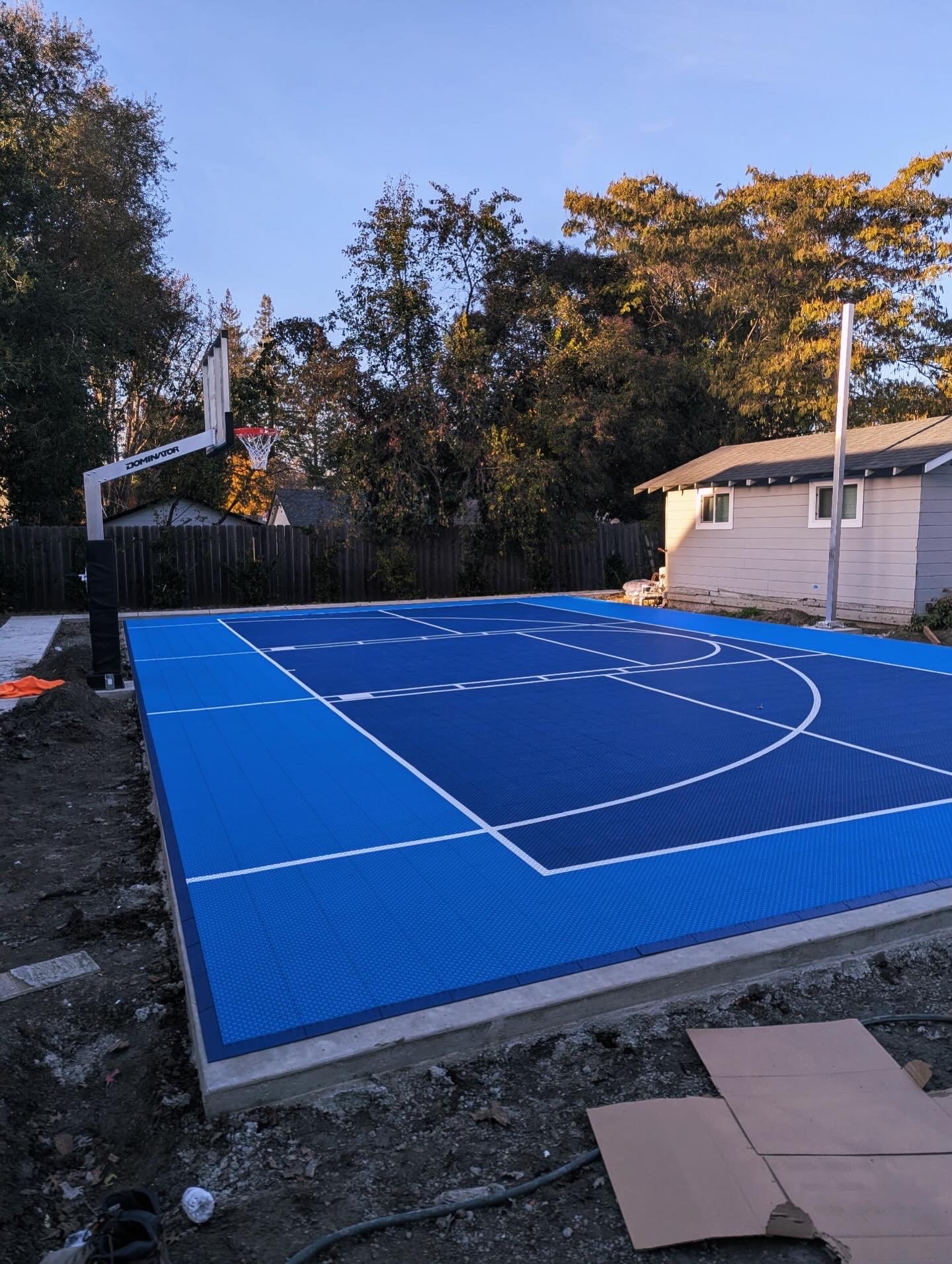 A newly installed outdoor blue basketball court with white lines, surrounded by a black fence, with trees and a house in the background, during the evening.