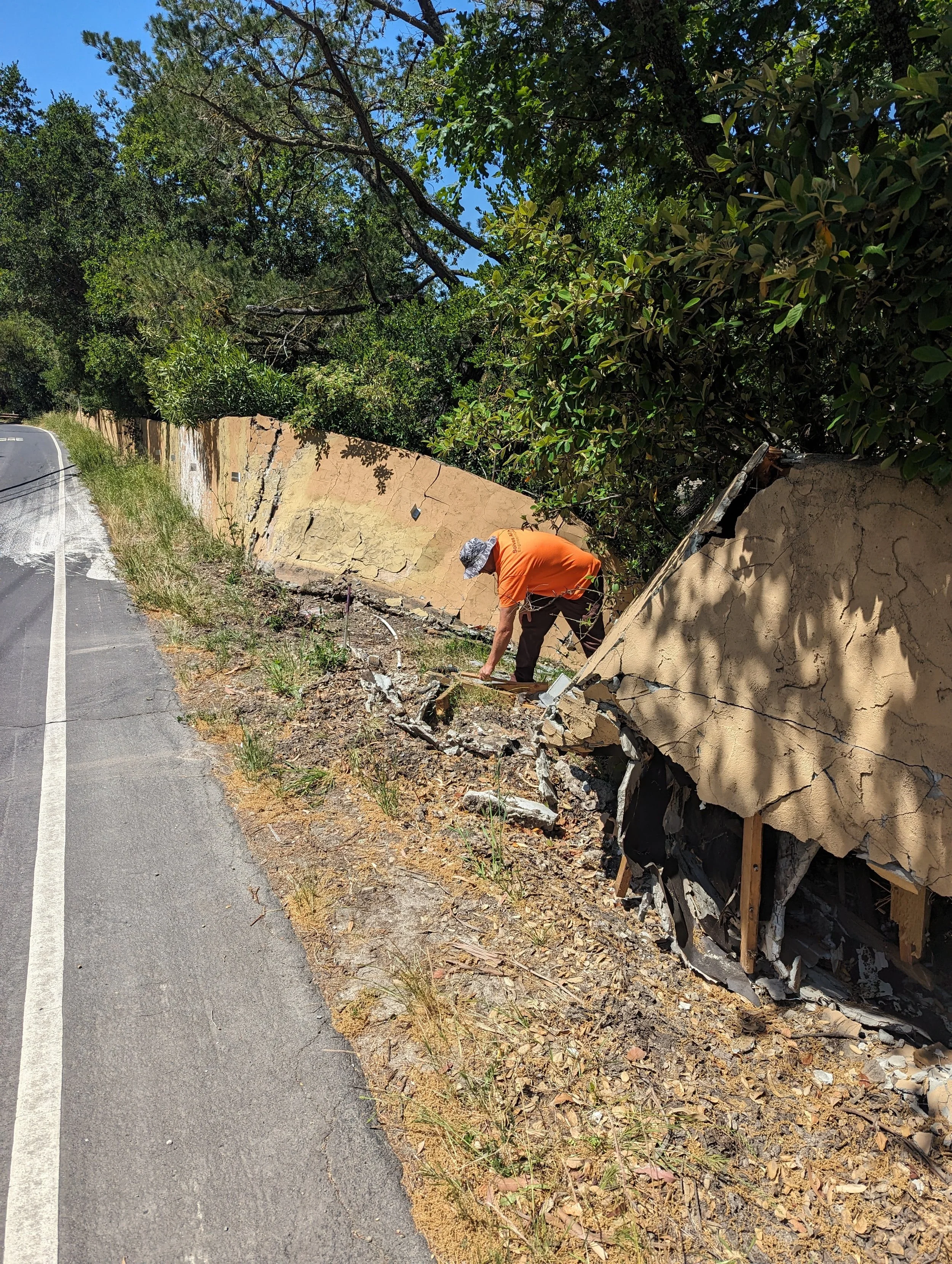 Person working to remove or assess damage to a cracked concrete and plaster retaining wall along the side of a road, with trees and clear blue sky in the background.