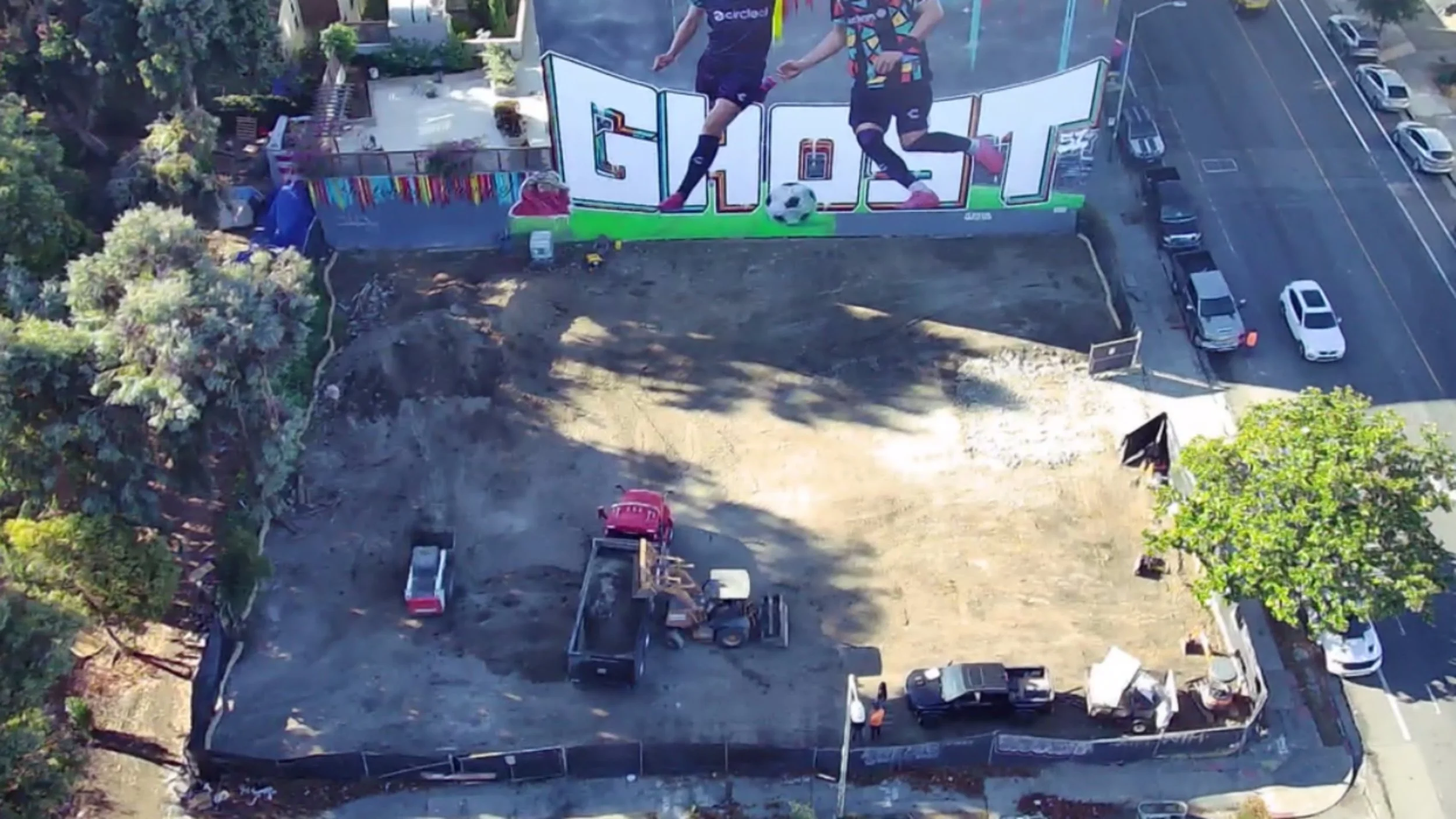 Empty construction lot with construction vehicles and equipment. A large mural of two soccer players kicking a ball is painted on a wall at the back of the lot. Surrounding the lot are trees, parked cars, and a sidewalk.