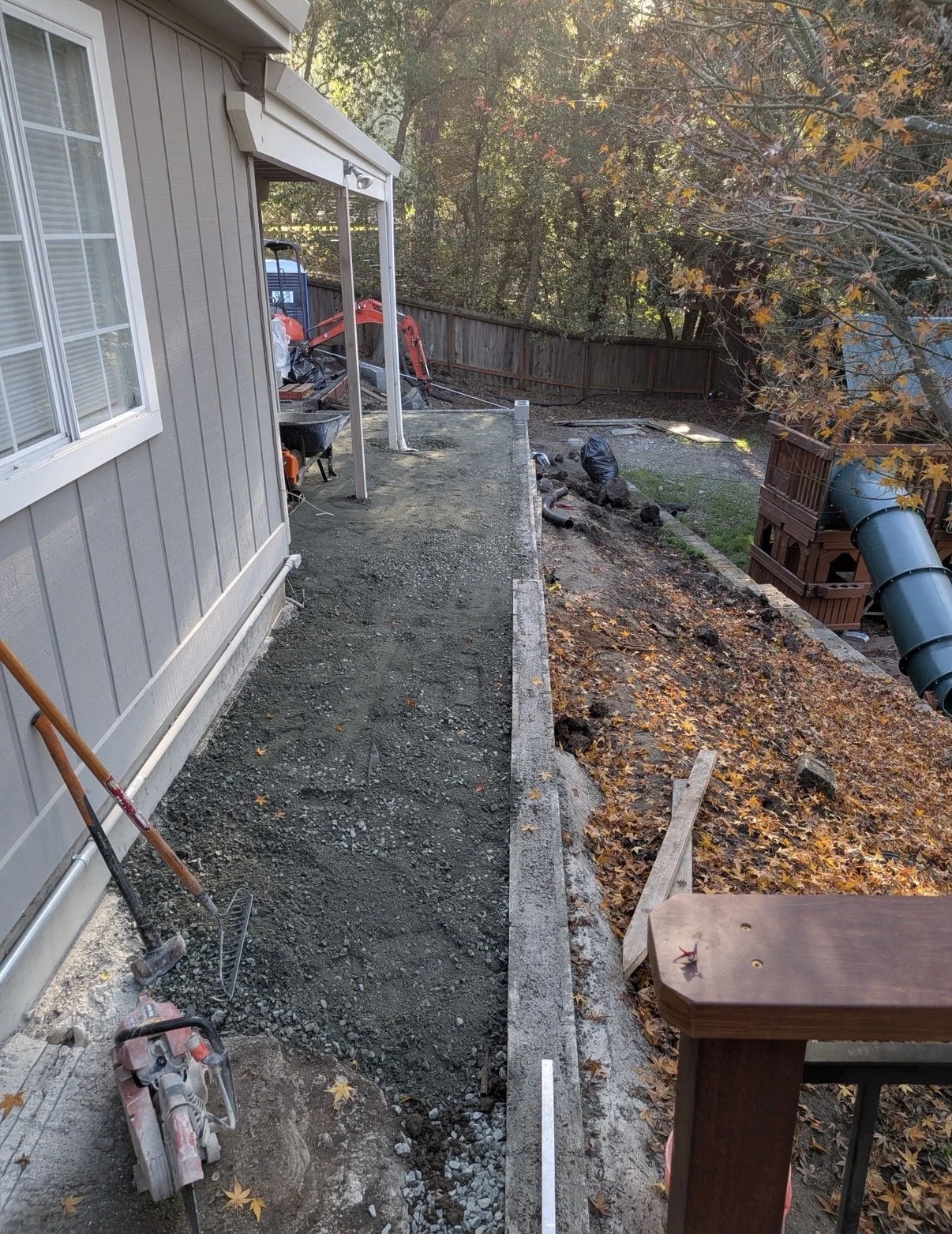 Backyard construction site with a new patio or walkway area being prepared with gravel, a wall in the process of being built, and driveway equipment. Leaves are scattered on the ground, and there is a green slide on the right side.