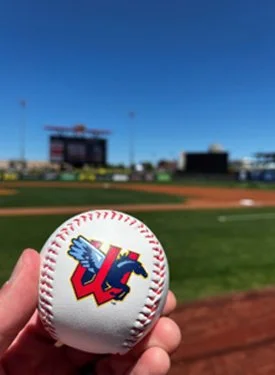 A hand holds a baseball featuring the Wichita Wind Surge logo in front of a blurry professional baseball field and scoreboard under a clear blue sky.