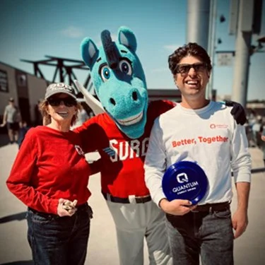 Two people, one in a "Better, Together" Quantum shirt holding a blue frisbee, pose with a blue horse mascot in a red "Surge" jersey at a stadium.