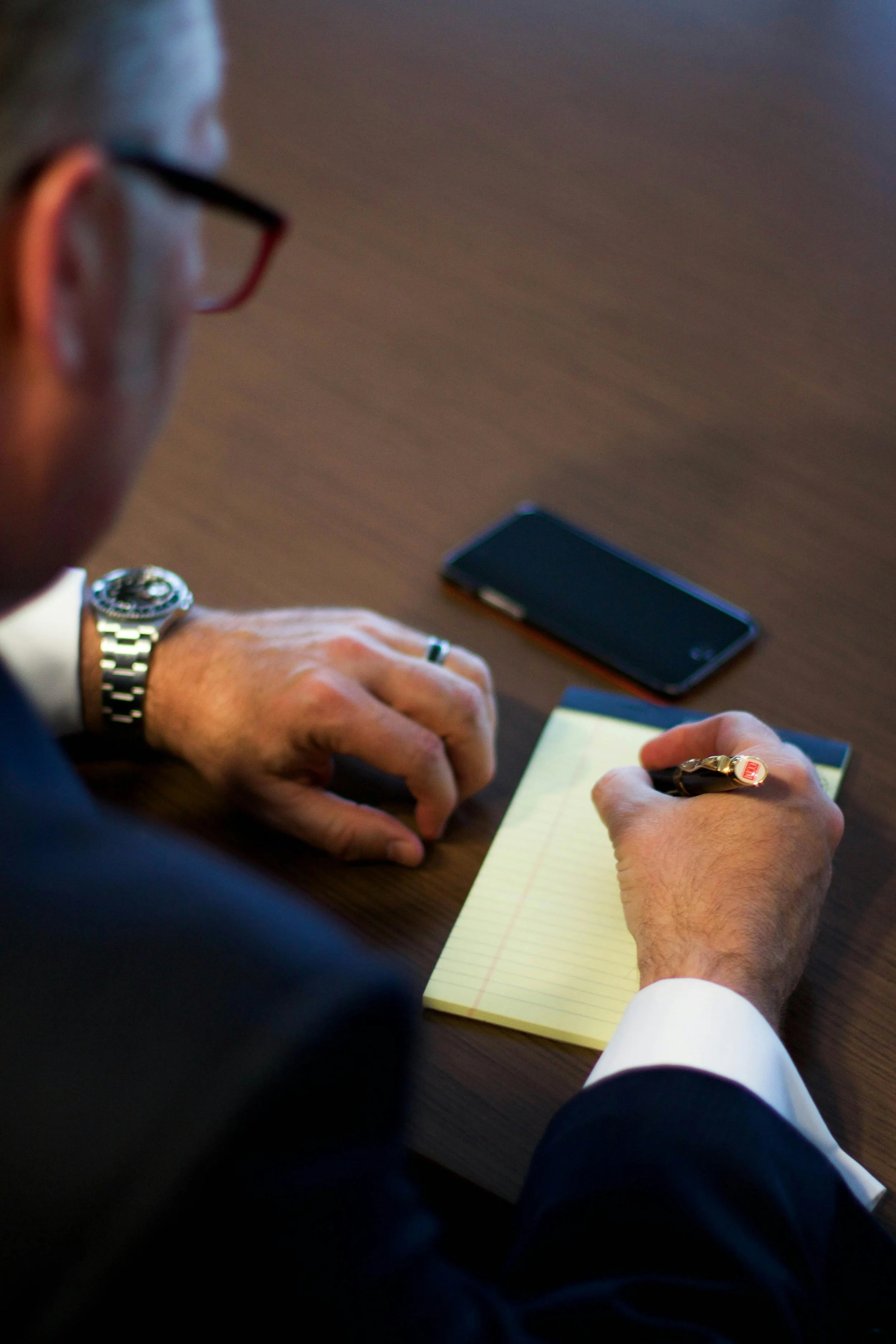 A man in a suit taking notes on a yellow notepad, with a smartphone on the table nearby.