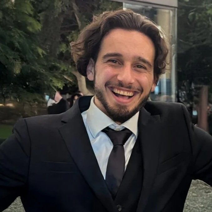 Timothy Grech wearing a black suit and tie, smiling outdoors with trees and glass building in the background.