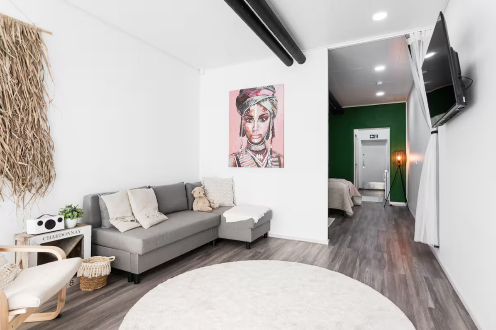 Living room with gray sofa, white wall art, and a TV on the wall, leading to a bedroom with green accent wall and a floor lamp.