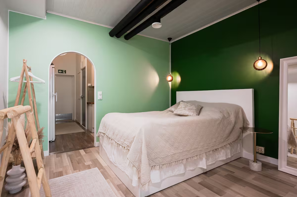 A bedroom with green walls, a white bed with beige bedding and a pillow, hanging pendant lights, a mirror, and a coat rack.