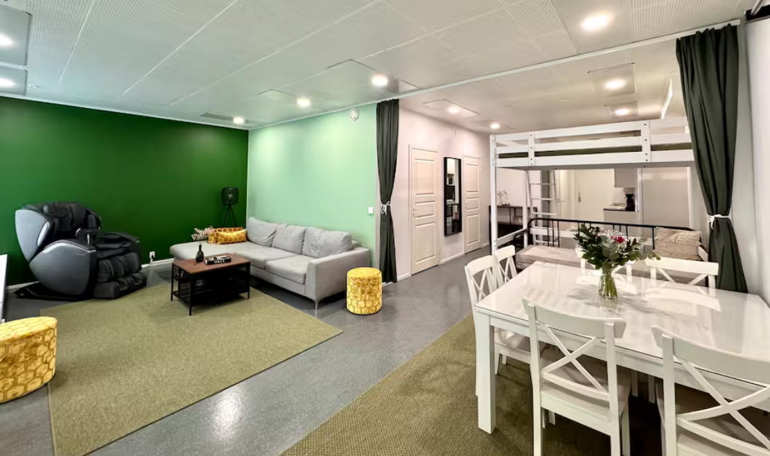 Living room with green accent wall, gray sofa, massage chair, yellow ottomans, and coffee table. Adjacent dining area with white table and chairs, and a loft bed with black curtains.