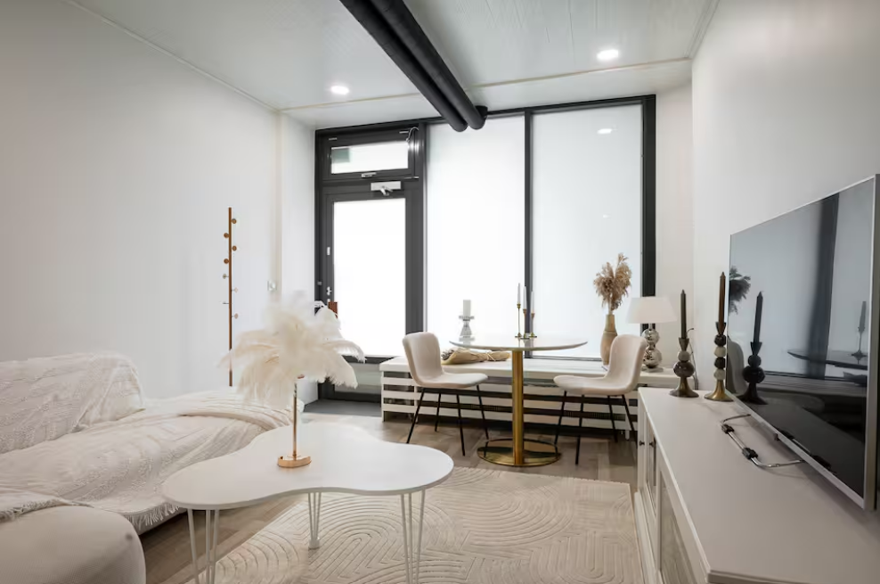 Modern living room with white walls, a small round dining table with two chairs, a TV on the right, and a white couch on the left. Decor includes candles, a lamp, and a vase with pampas grass, with large windows in the background.