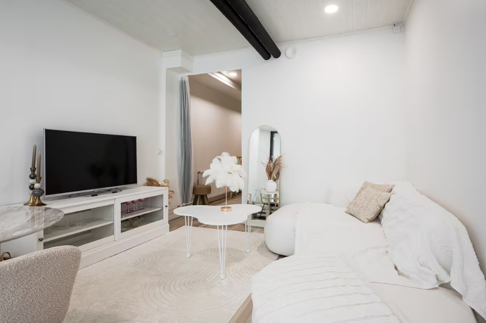 Bright living room with white walls and modern decor, featuring a flat-screen TV on a white console, a stylish round white coffee table with a feather centerpiece, a white upholstered sofa with beige pillows, and a small mirror with dried flowers behind it.