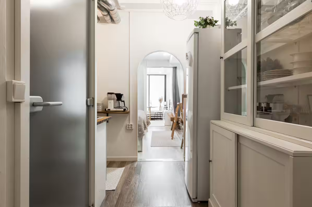 A narrow kitchen view toward a dining area with a large window and archway, white cabinets, and wooden floors.