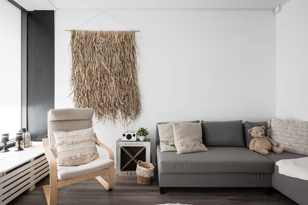 Modern living room corner with a beige armchair, gray sectional sofa, wall hanging made of dried grass, small side table with decorative items, teddy bear on the sofa, and neutral-colored cushions.