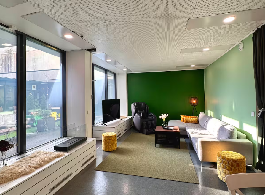 A modern living room with large floor-to-ceiling windows, a white sectional sofa with yellow accent pillows, a black massage chair, a small black coffee table, a green accent wall, and a yellow ottoman. There's also a tall black floor lamp, a vase with flowers, and a TV on a white media console. The room has a beige rug and a view of an outdoor patio through the windows.