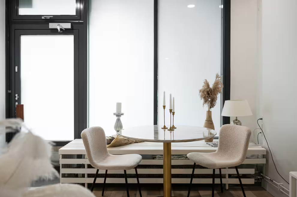 Interior of a modern room with a round dining table, two beige upholstered chairs, a window with black frames, a white lamp, a vase with dried plants, and gold candlesticks.