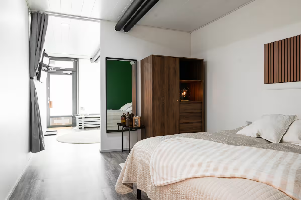 A modern bedroom with a bed, wooden wardrobe, wall decoration, and a mirror reflecting the bed and green wall. A door leads to a bright outdoor area with a white circular rug and a wall-mounted TV.
