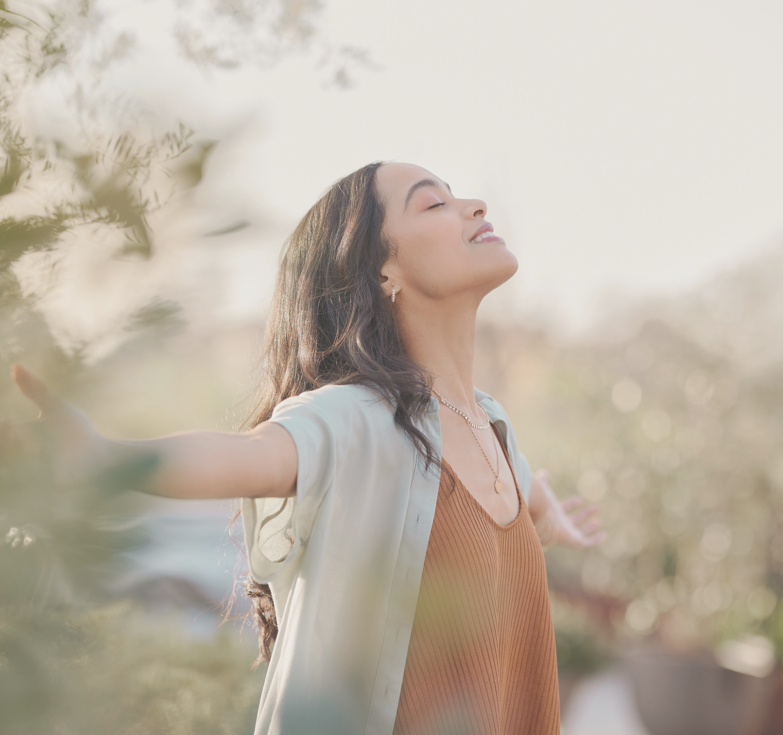 A woman standing outdoors with her arms outstretched, eyes closed, and a content smile, embracing sunshine and nature.